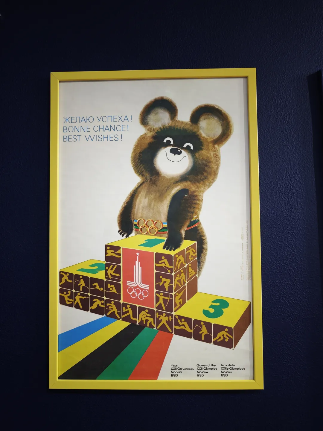 A poster with a yellow frame hanging on the wall shows a bear standing on a block. The bear is wearing an Olympic ring belt, and the background is white. Text in multiple languages appears below the poster.