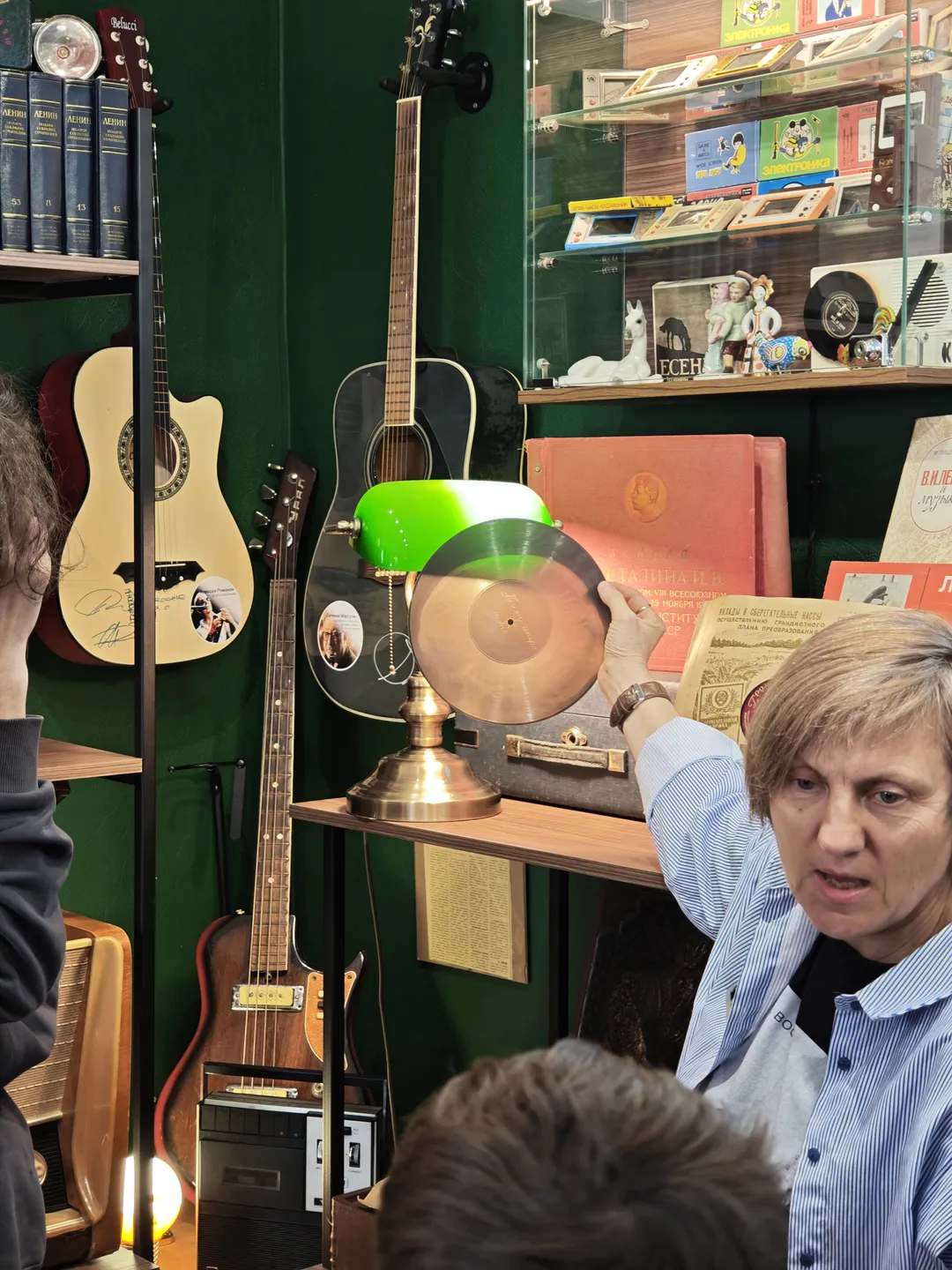 A person in a striped shirt is showcasing a golden record, with a green desk lamp beside them. Guitars and other musical instruments are visible in the background, and a display case holds various items.