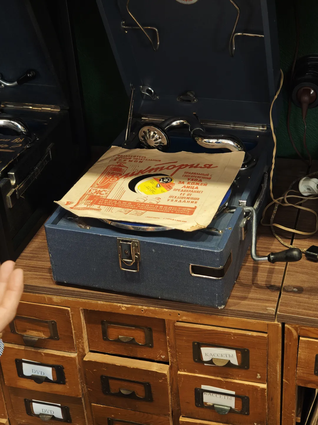 A blue vintage turntable placed on a wooden cabinet, with several drawers. A record and a piece of paper with Russian text are on top of the turntable. Nearby, there are wires and headphones.