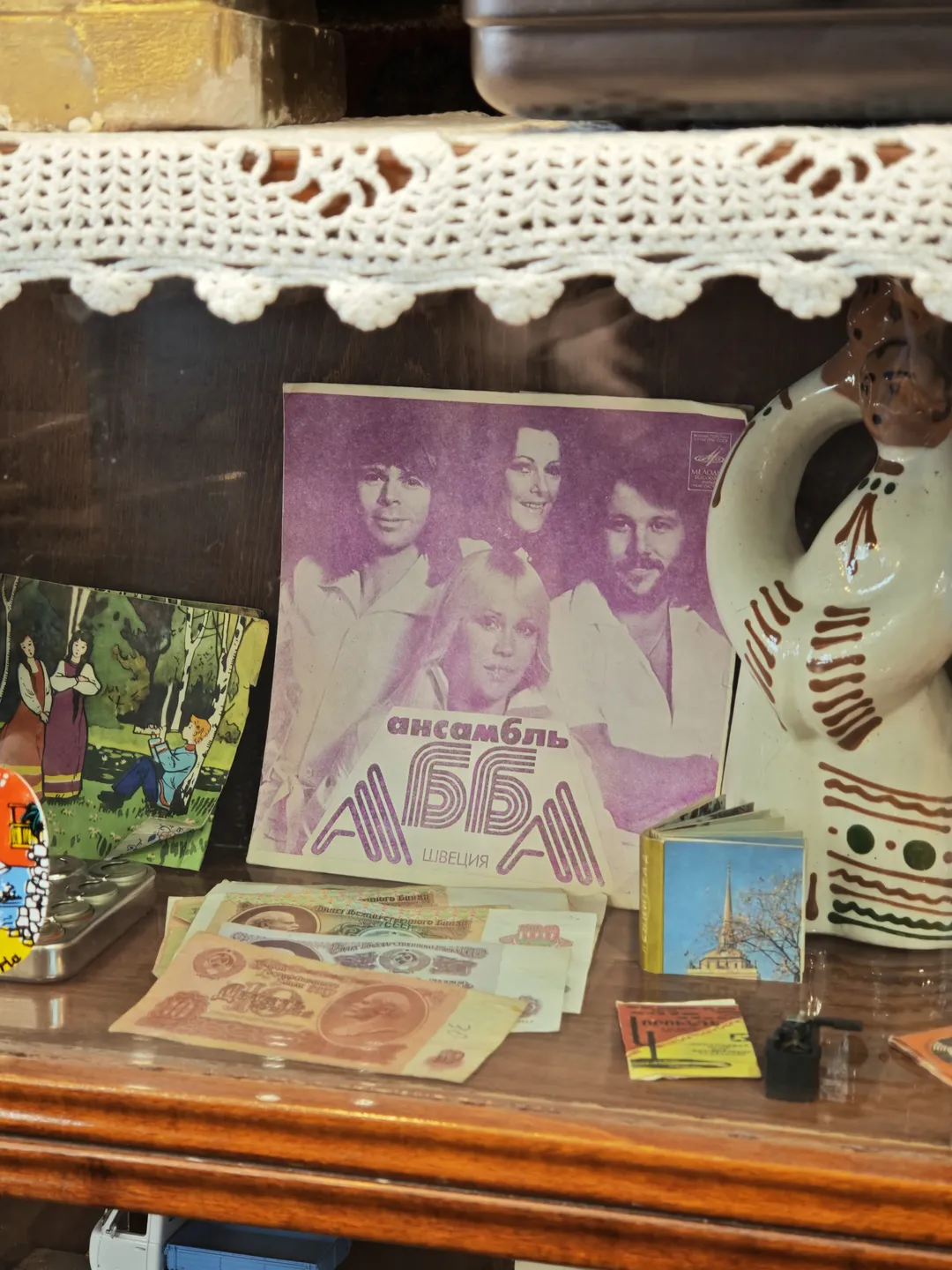 A display case filled with various items, including a book with a purple cover, some paper money, and a small ceramic pot. The book’s cover features images of people, and the background is purple. Above the cabinet, there is a white lace cloth.