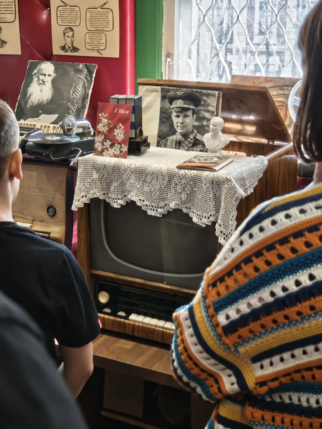 Two people stand in front of an old-fashioned television, which is covered with a white lace cloth. Above the TV, two black-and-white photos are hung, and a small sculpture sits nearby. The background wall is red, with posters attached.