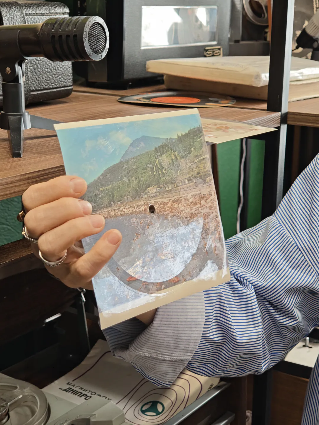 A person holds a transparent film with a landscape painting on it. The edges of the film have holes, and in the background, some recording equipment is visible. The person is wearing a striped shirt and a ring on their finger.