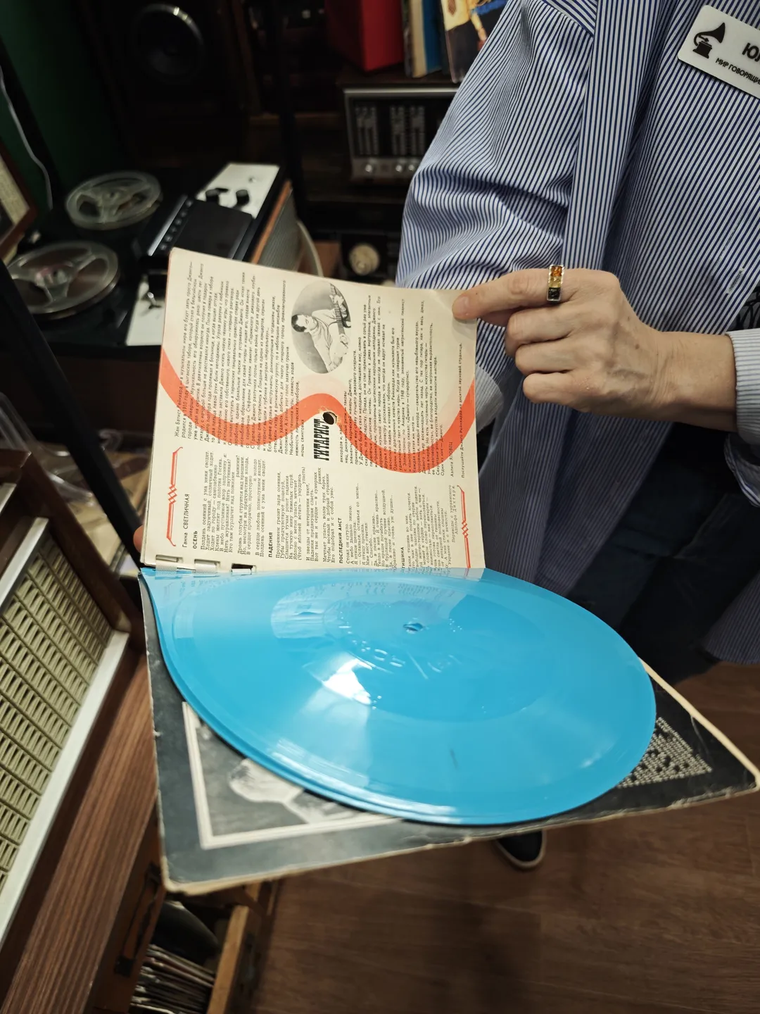 A person is holding a blue record, which is placed in an open sleeve. The inner side of the sleeve features text and patterns. Electronic equipment is visible in the background. The person is wearing a striped shirt and a ring on their finger.