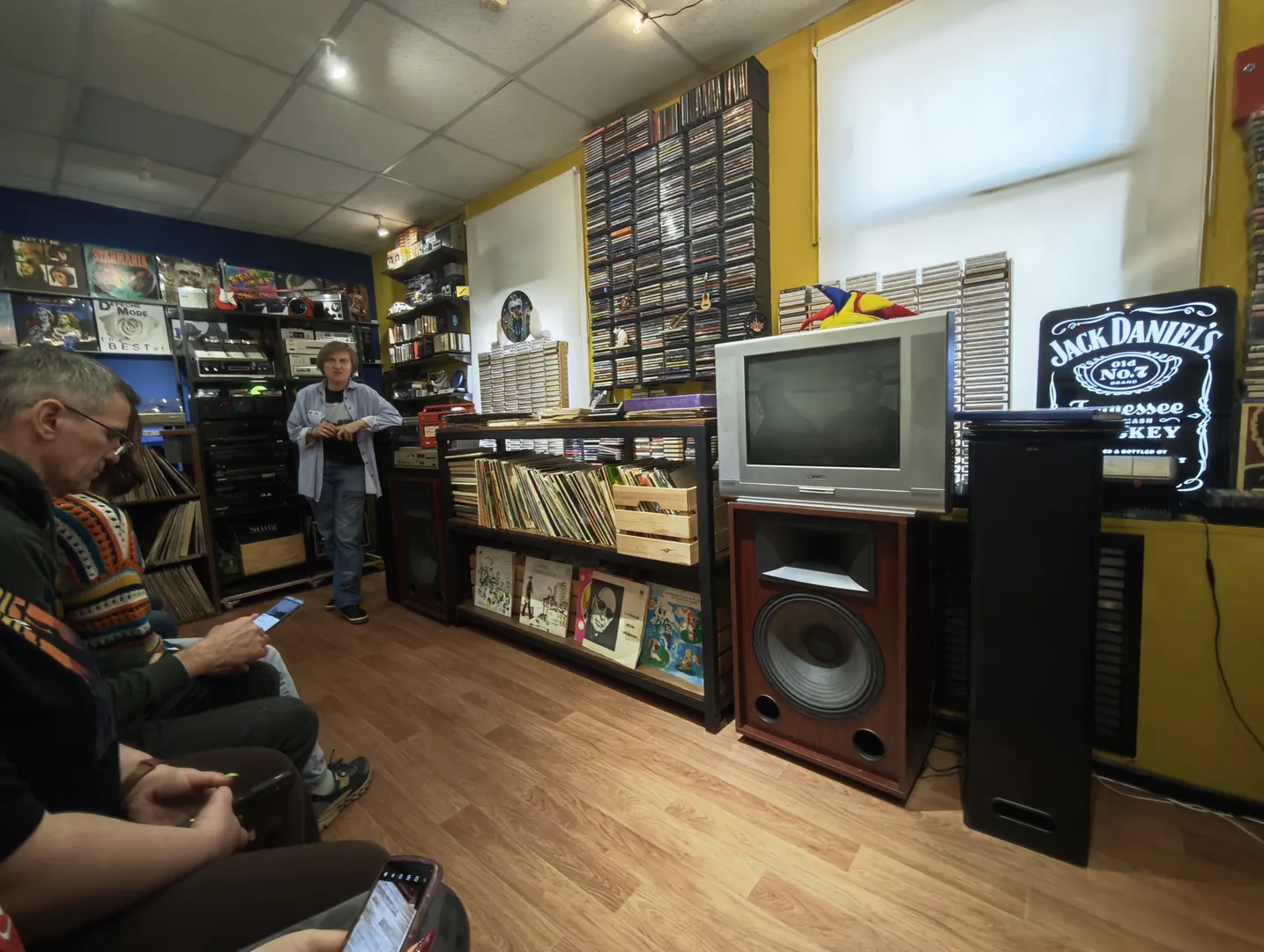 In a room with several people, one person is sitting on a sofa using their phone. The room is filled with record shelves and sound equipment, with multiple screens on the wall. Another person stands in front of the record shelf, holding an item.