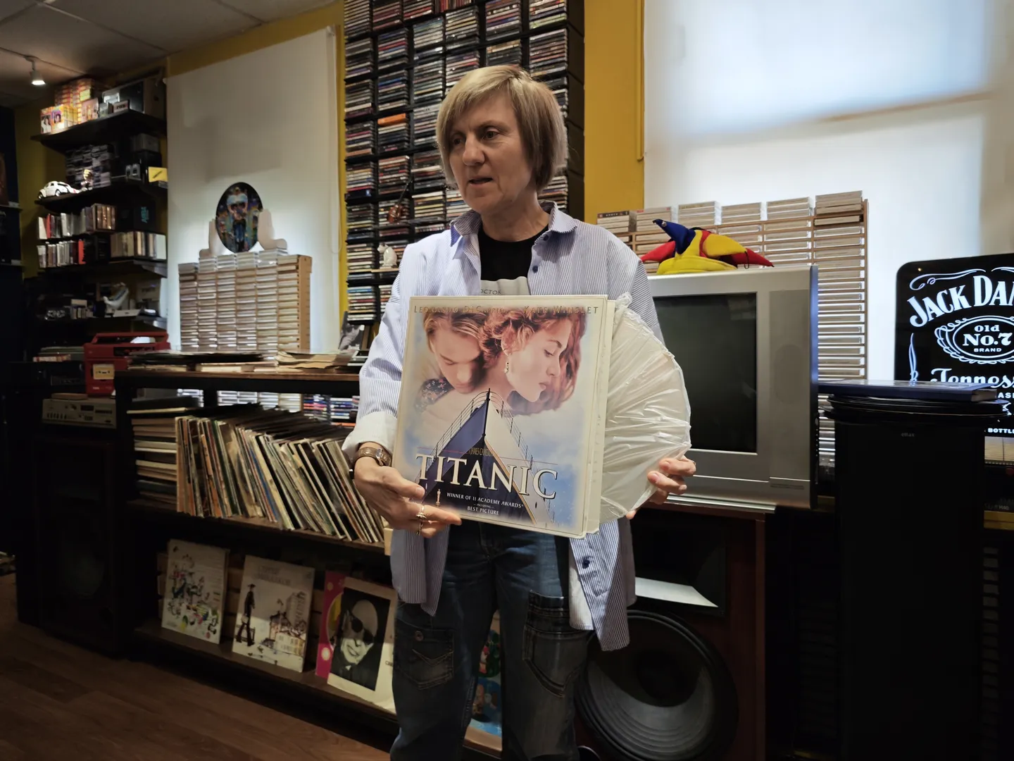 A person is holding a movie poster for Titanic, featuring the images of the lead male and female characters. The person, wearing a striped shirt, stands in front of a record shelf. In the background, several records and sound equipment are visible.