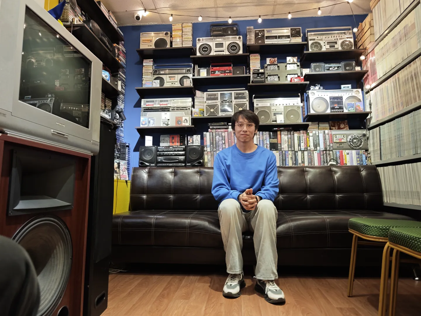 A person is sitting on a black leather sofa. Behind the sofa are multiple shelves displaying various sound equipment. The person is wearing a blue top and light-colored pants, with their hands crossed on their knees. The room’s walls are blue, decorated with more sound equipment.