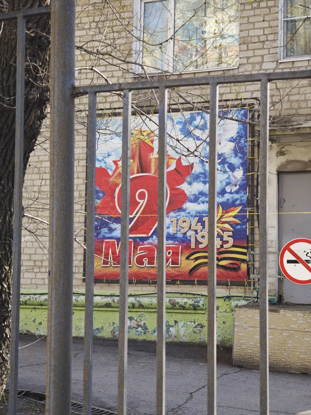 On a wall, a large poster featuring the words “1941-1945” and related images is displayed. In front of the poster is a metal railing, behind which a tree is visible. The building’s exterior wall is made of light-colored bricks, and the windows reflect the surrounding scene.