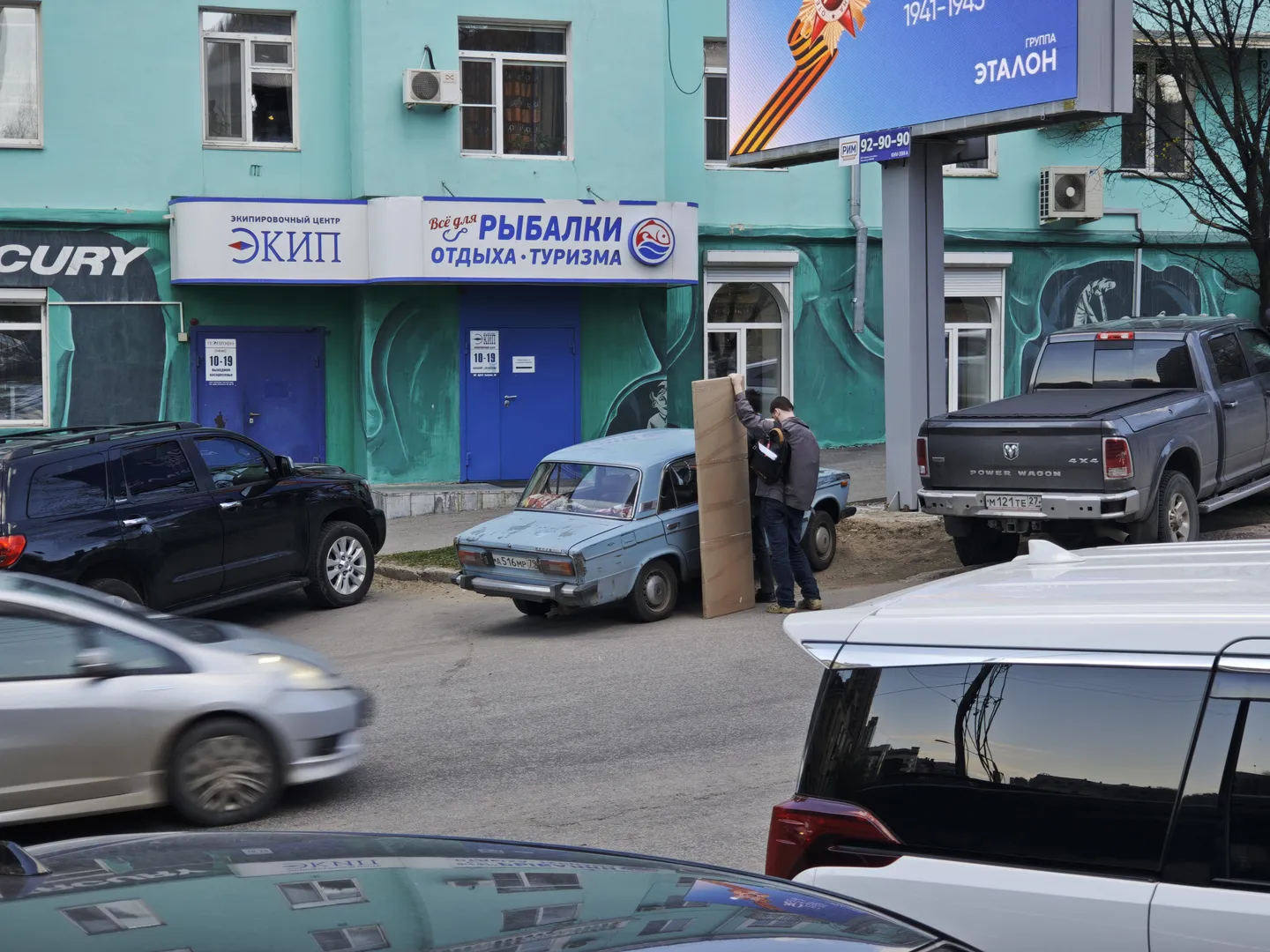 On the street, several cars are parked, one of which is an old vehicle. A person is unloading a large cardboard box from the car. The building’s exterior is light blue, and the door features the text “РЫБАЛКИ ОТДЫХА ТУРИЗМА.” Above the street, a large billboard displays “1941-1945” and related imagery.