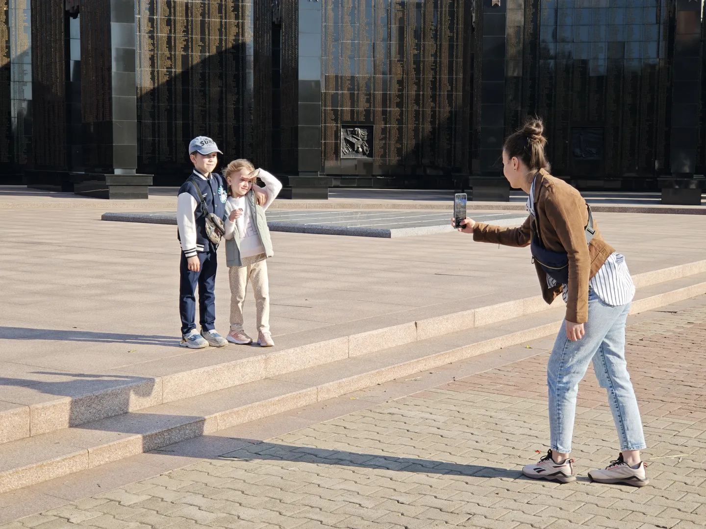 A woman is taking a photo of two children standing on stairs. The background features a large building.