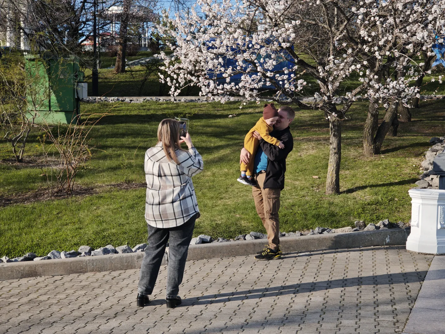 A man is holding a child, while a woman is taking a photo. In the background, trees in full bloom and a grassy field are visible.