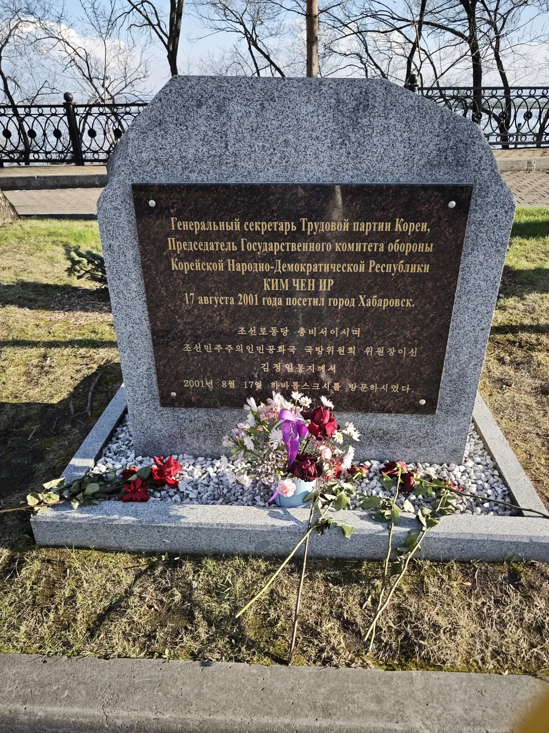 A stone tablet is inscribed with Russian and Korean text. In front of it, some flowers and purple ribbons are placed.