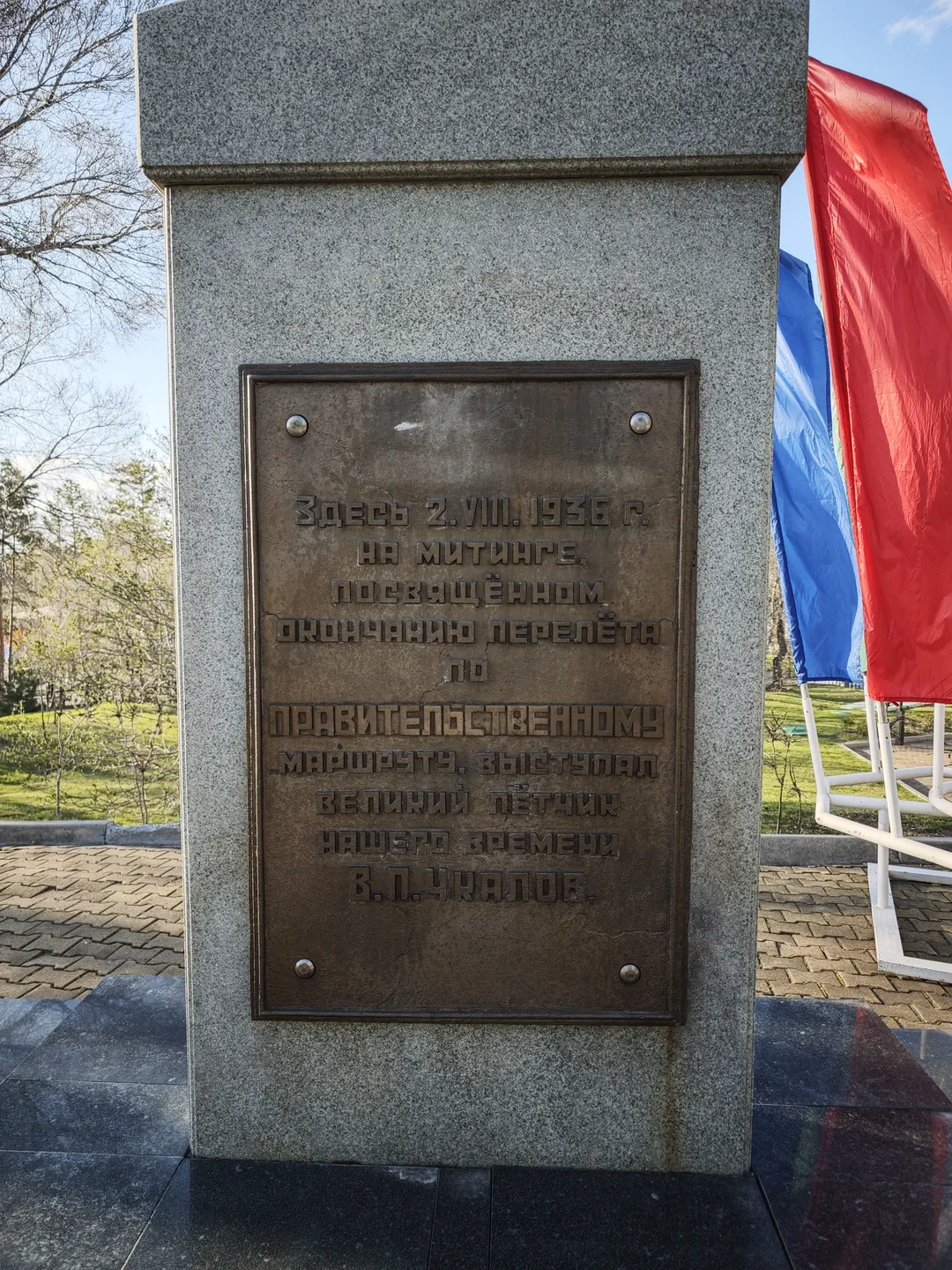 A stone tablet is inscribed with Russian text. Nearby, two flags are displayed, and the background features trees and grass.