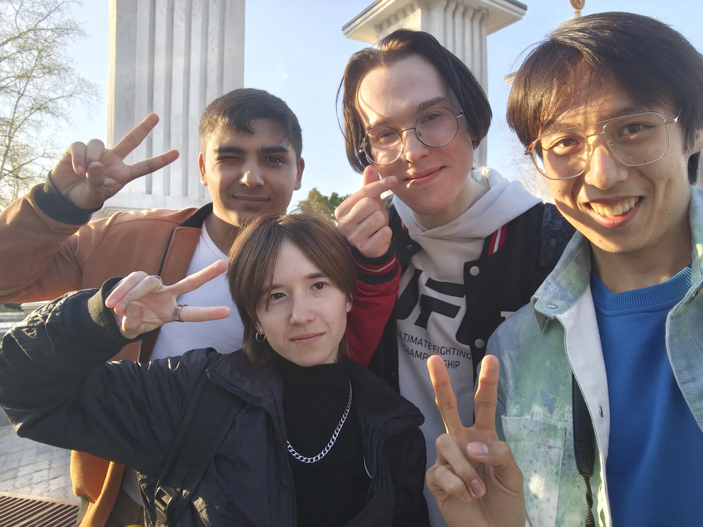 Four people stand together for a group photo, with three of them making victory signs. The background features columns and the sky.