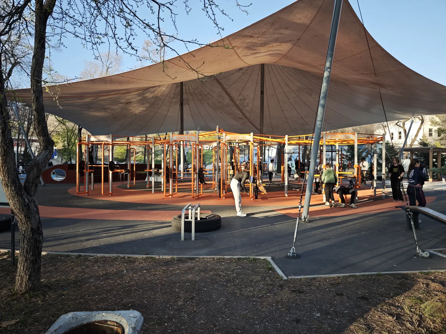 An outdoor fitness area with a sunshade on top, where several people are using the gym equipment.