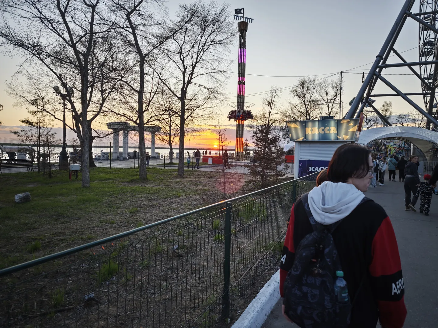 A person carrying a backpack walks along a road. Ahead, there is an amusement facility, and the background features the sunset and trees.
