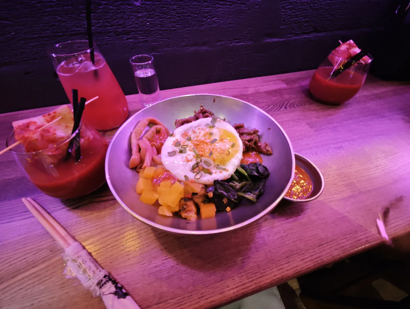 A bowl of food on a table, including fried eggs, vegetables, and meat. Several drinks are placed beside it, one of which has a straw. A pair of chopsticks is positioned in the lower left corner of the table.