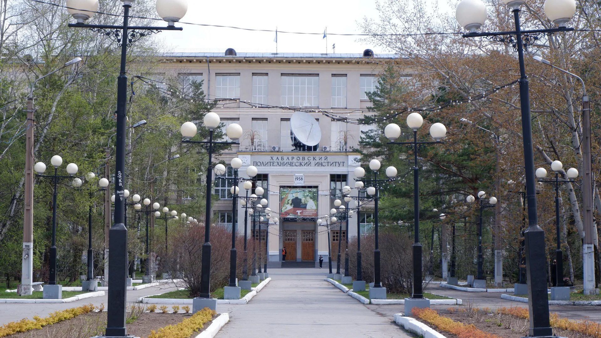 A wide road with streetlights leads to a building marked “ХАБАРОВСКИЙ ПОЛИТЕХНИЧЕСКИЙ ИНСТИТУТ,” with a large satellite antenna in front of it. Trees and shrubs line both sides of the road. There are several wooden doors at the entrance of the building.