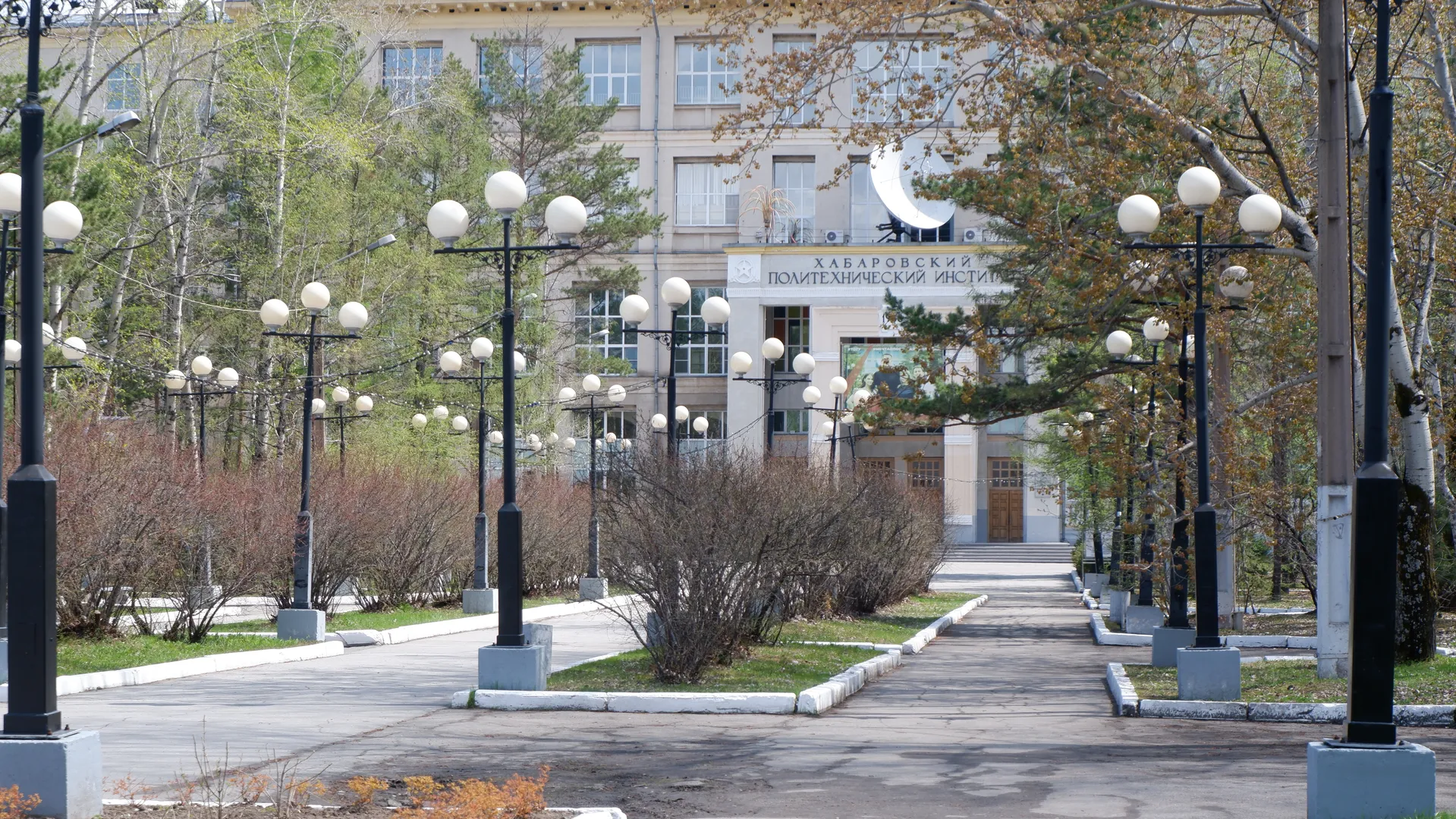 A street lined with streetlights leads to a building marked “ХАБАРОВСКИЙ ПОЛИТЕХНИЧЕСКИЙ ИНСТИТУТ,” with a large satellite antenna in front of it. Trees and shrubs line both sides of the road. Several wooden doors are at the entrance of the building.