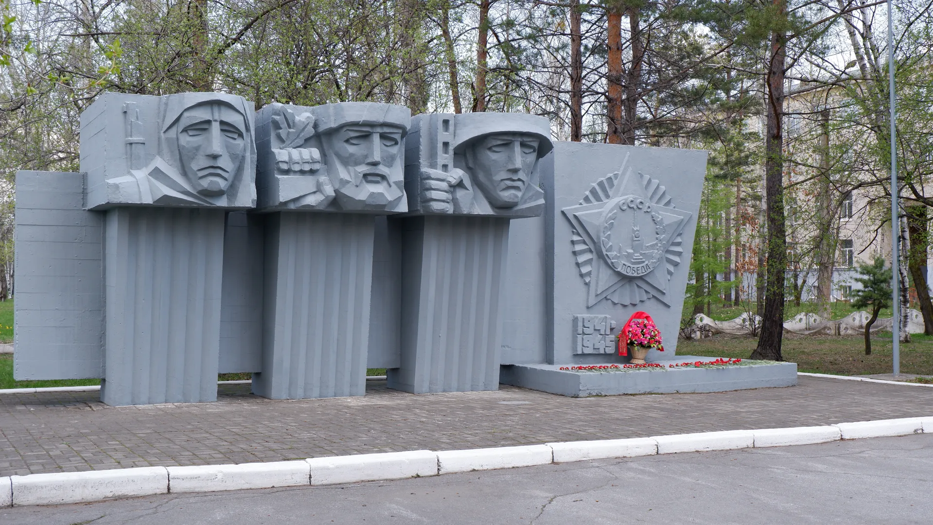 A gray monument features three sculpted faces and a Soviet emblem, with the inscription “1941-1945.” A bouquet of flowers is placed in front of the monument. Trees and some buildings can be seen in the background.