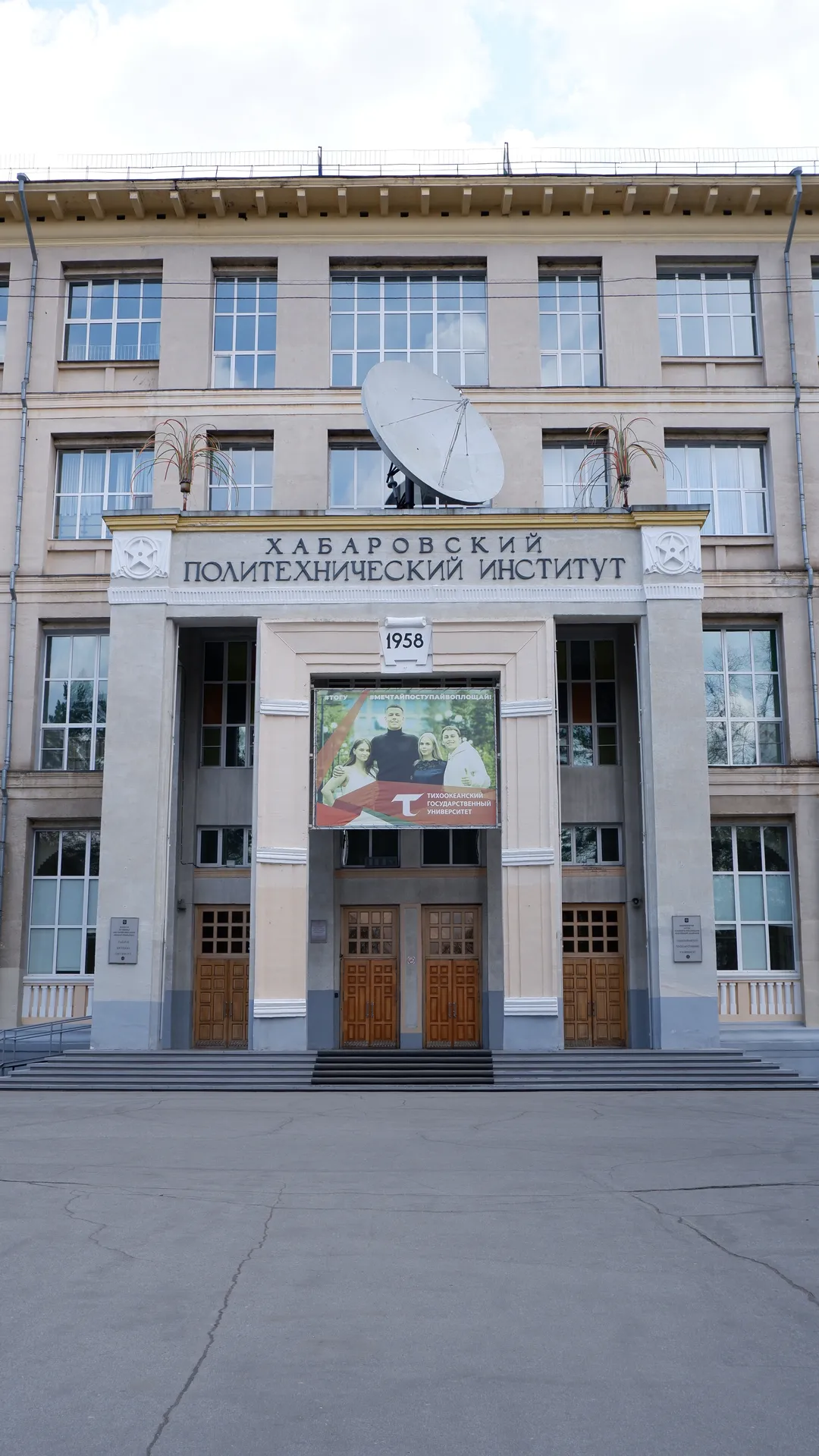 A building marked “ХАБАРОВСКИЙ ПОЛИТЕХНИЧЕСКИЙ ИНСТИТУТ” has a large satellite antenna in front of it. Several wooden doors are at the entrance of the building. Above the doors, a poster shows images of several people.