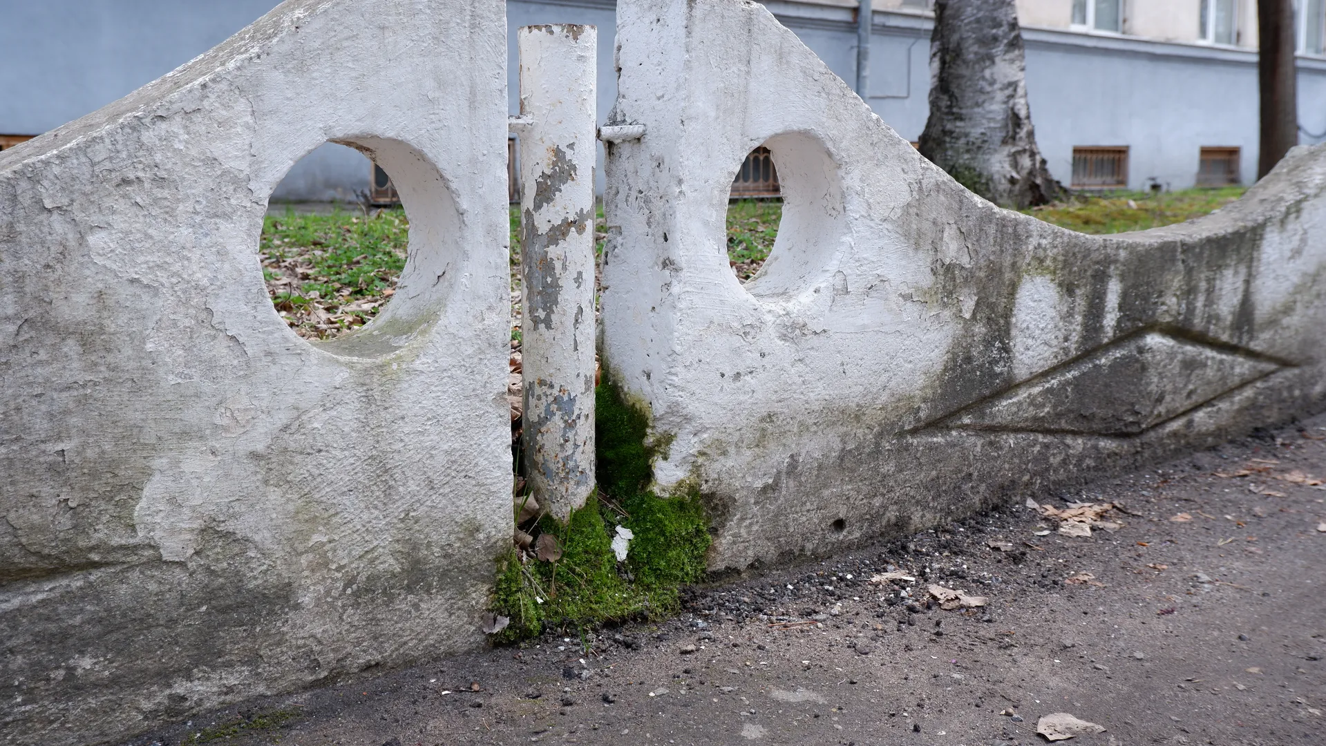 A white stone fence with round holes, its surface showing peeling paint. Some green moss can be seen at the bottom of the fence. In the background, there are some buildings and trees.