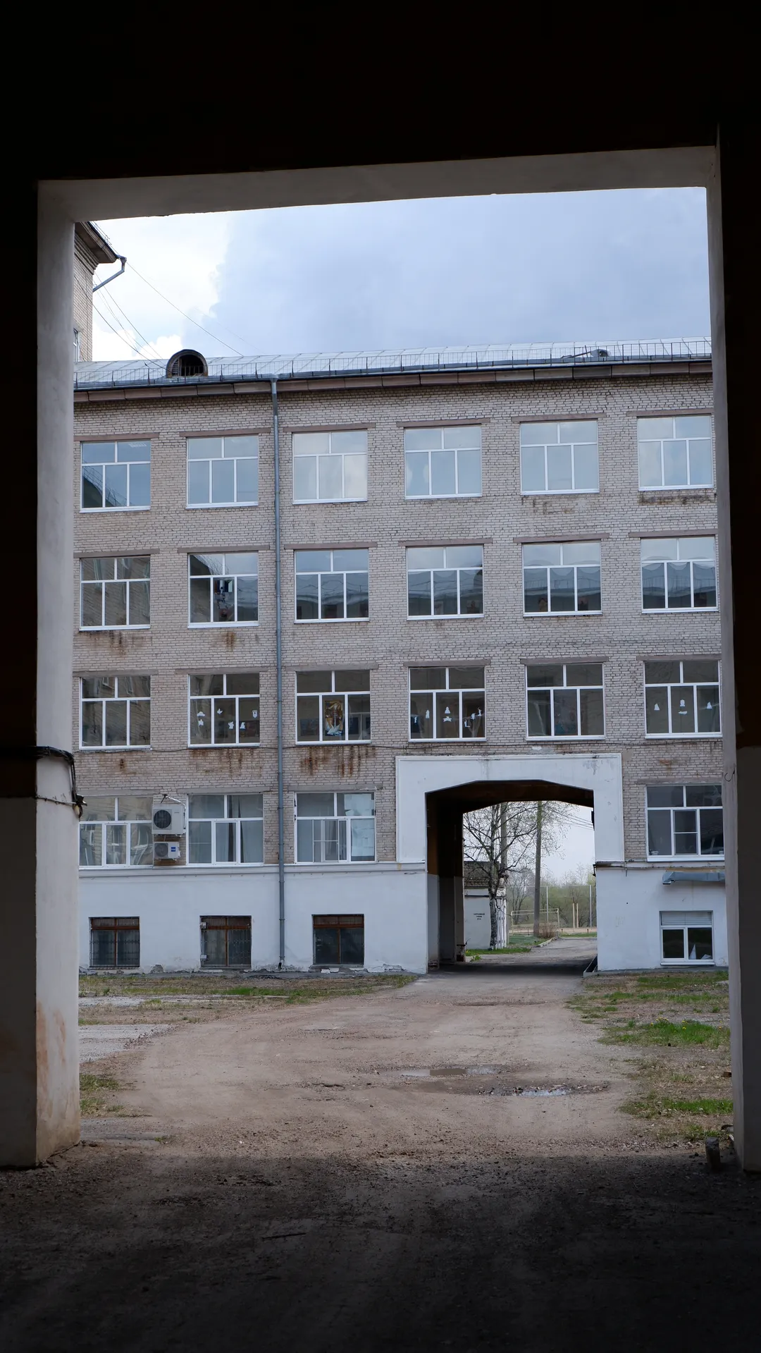 Looking through an archway, a multi-story brick building with many windows is visible. The lower level has an arched doorway leading into the distance. The sky is cloudy.
