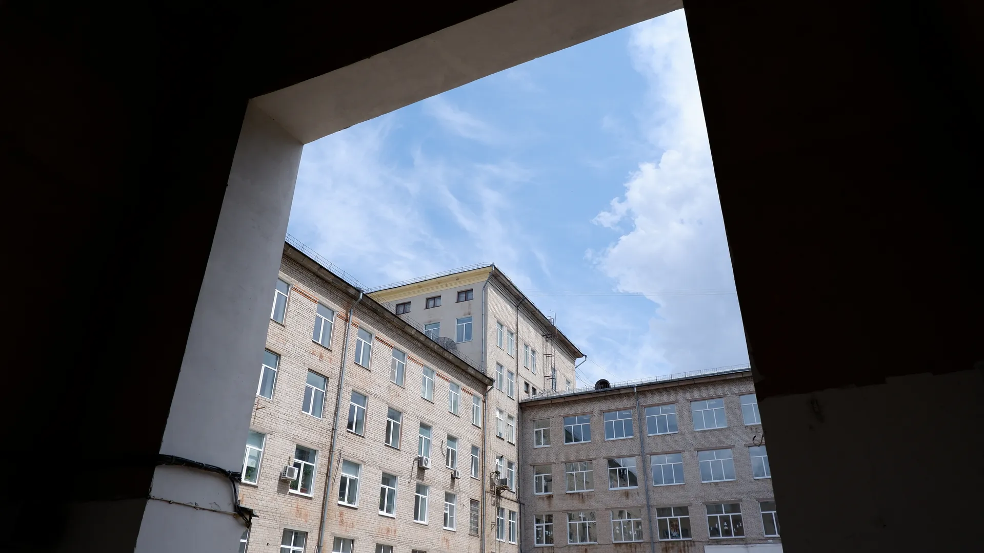 Looking through an archway, a multi-story brick building with many windows is visible. The side of the building has pipes and air conditioning units. The sky is cloudy.