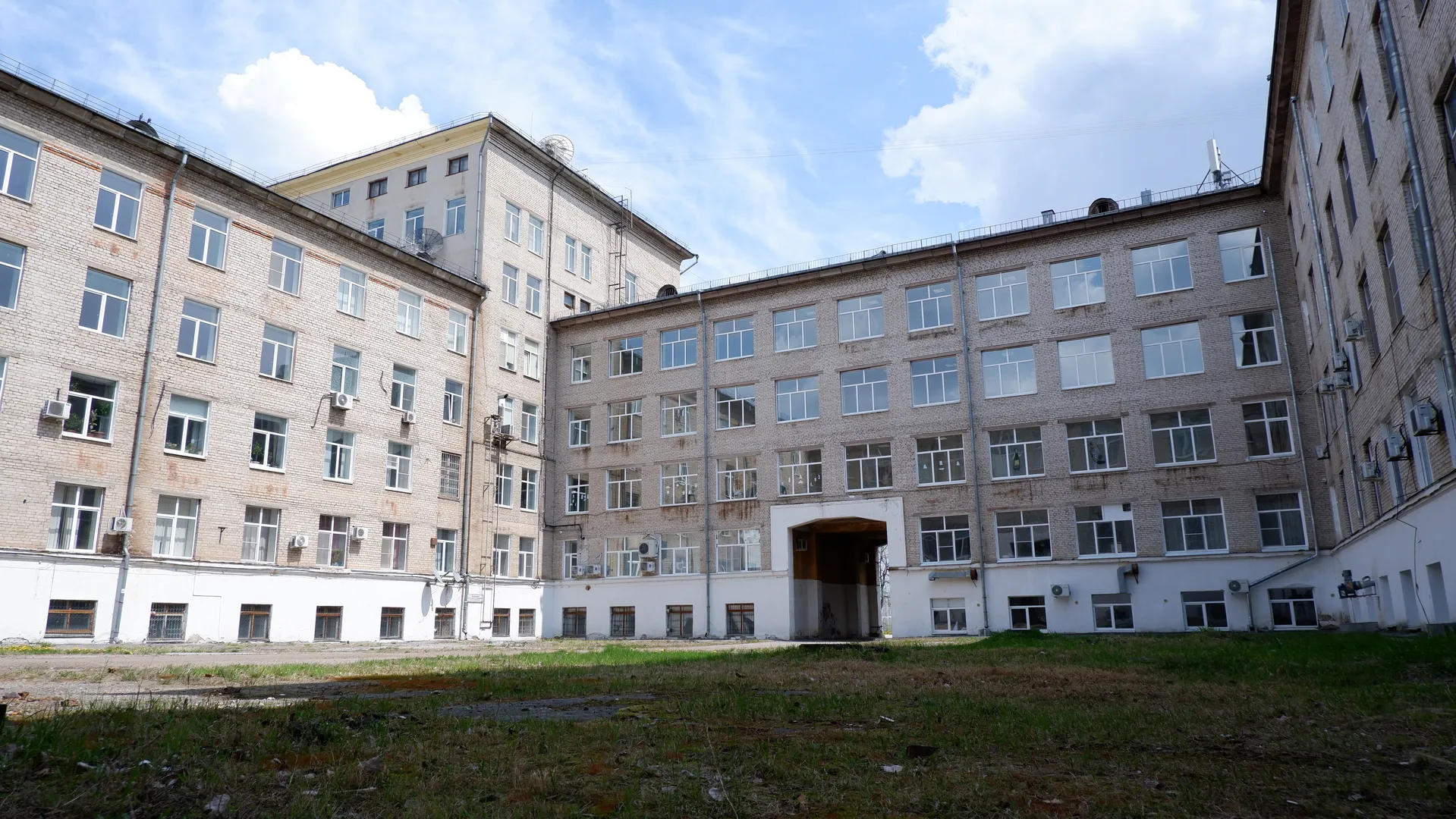 A multi-story building with light-colored brick walls and neatly arranged windows. There is a patch of grass in front of the building, and a few white clouds in the sky. Several satellite antennas are installed on the roof of the building.