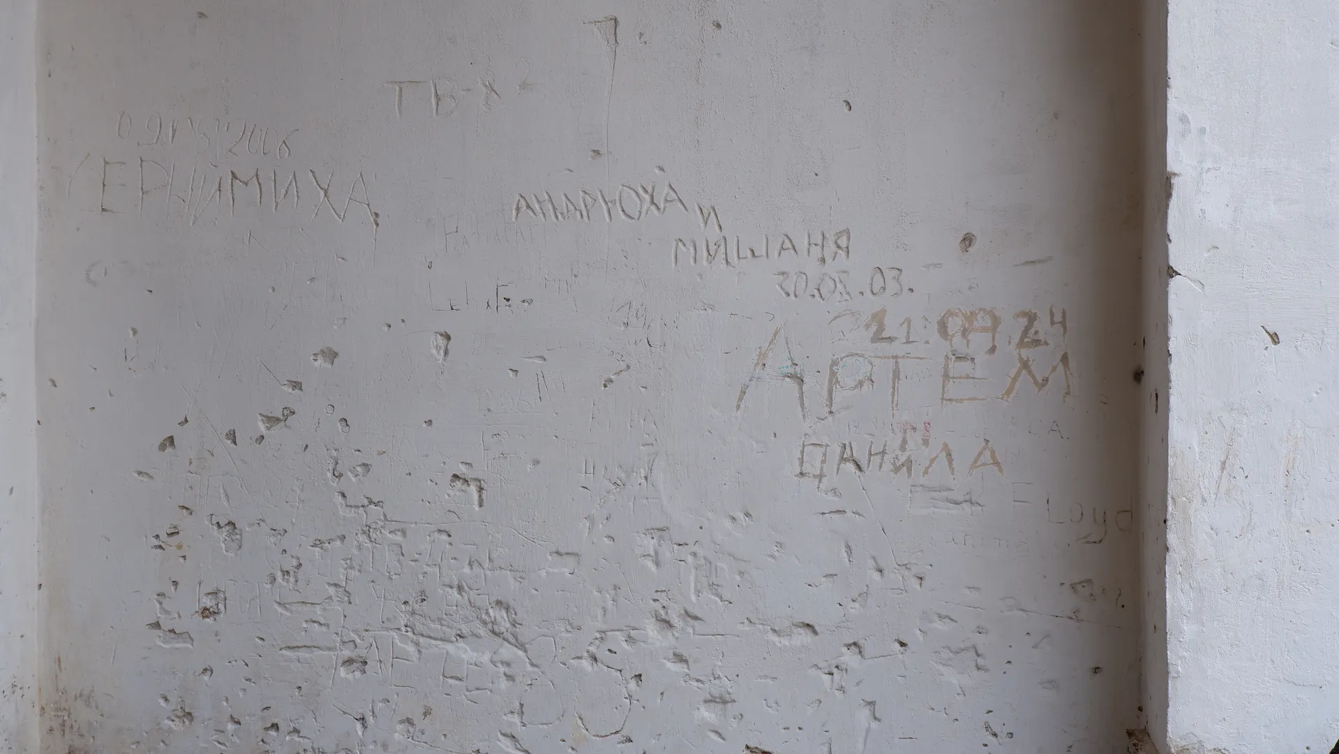 A white wall is engraved with Russian names and dates, such as “Артем” and “30.08.03.” The surface of the wall is rough, with some peeling marks. There are also other unclear carvings on the wall.