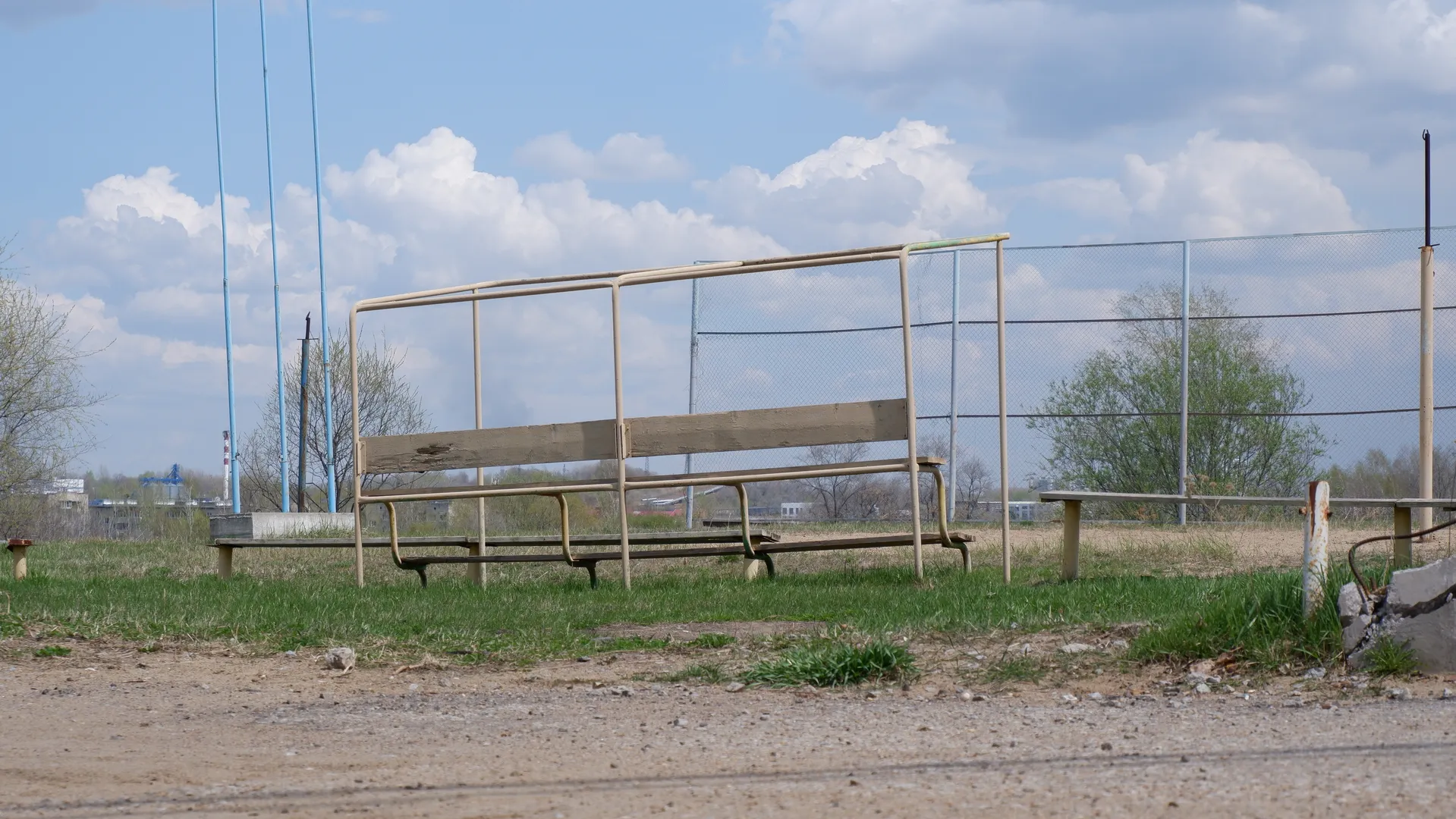 An open space with several old benches in the foreground. Behind the benches is a meadow and a wire fence. In the distance, some power poles and buildings can be seen.