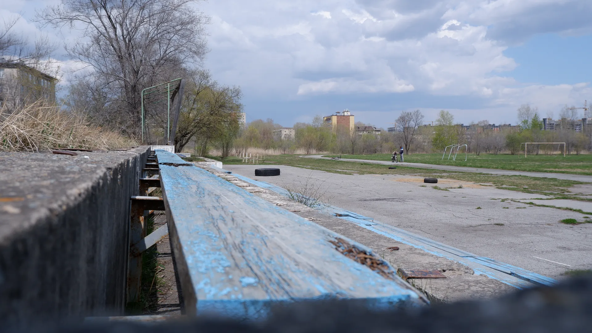 A blue bench with peeling paint placed in an open space. Behind the bench is a spacious area scattered with tires. In the distance, trees and buildings can be seen.