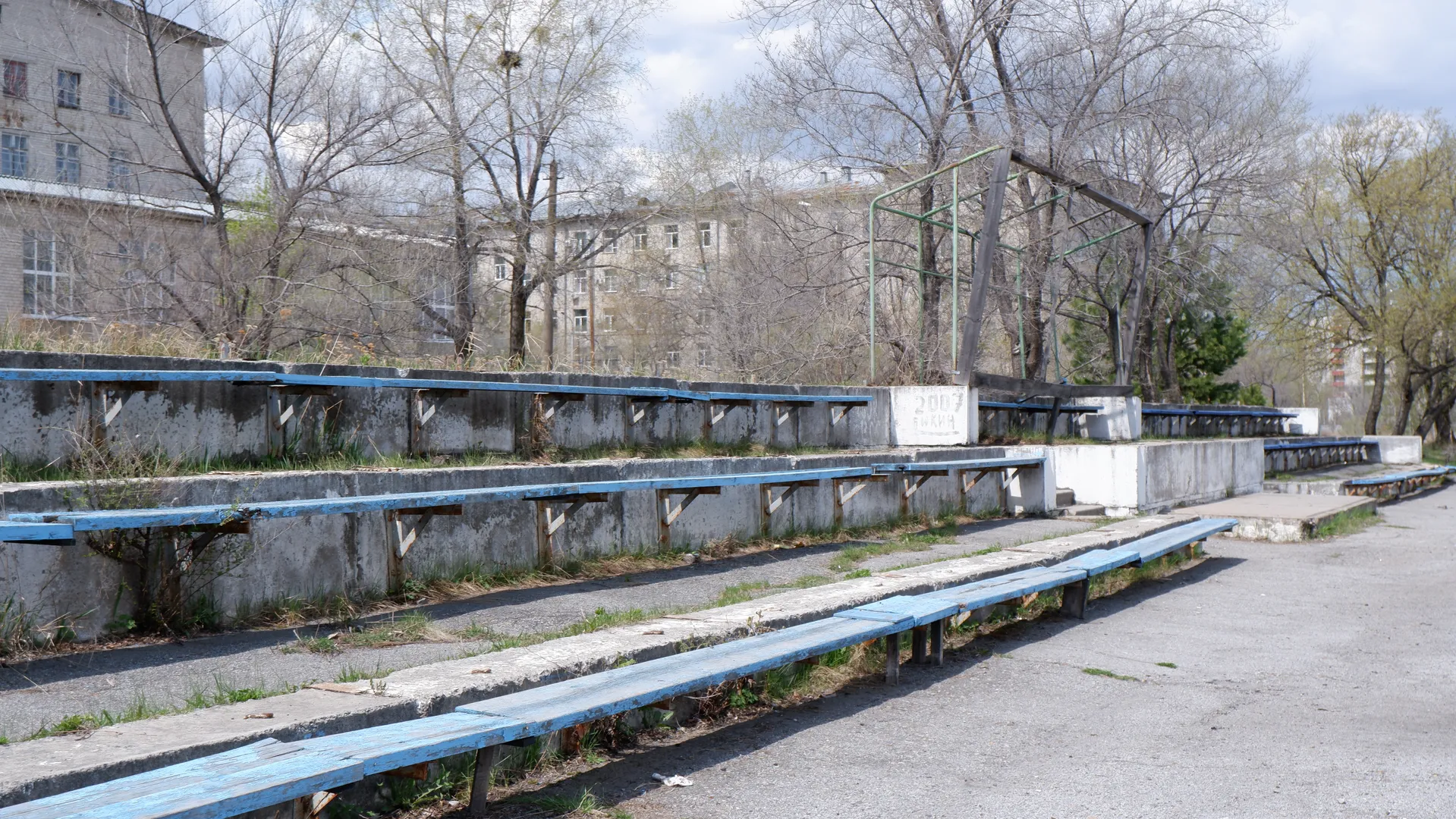A row of blue benches arranged on a gray surface. Behind the benches are stairs and more benches. In the background, trees and buildings can be seen.