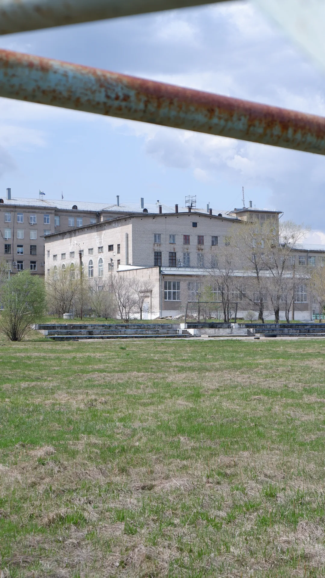 A meadow with a rusted metal frame in the foreground. Behind the meadow is a multi-story building with many windows. In the distance, trees and power poles can be seen.