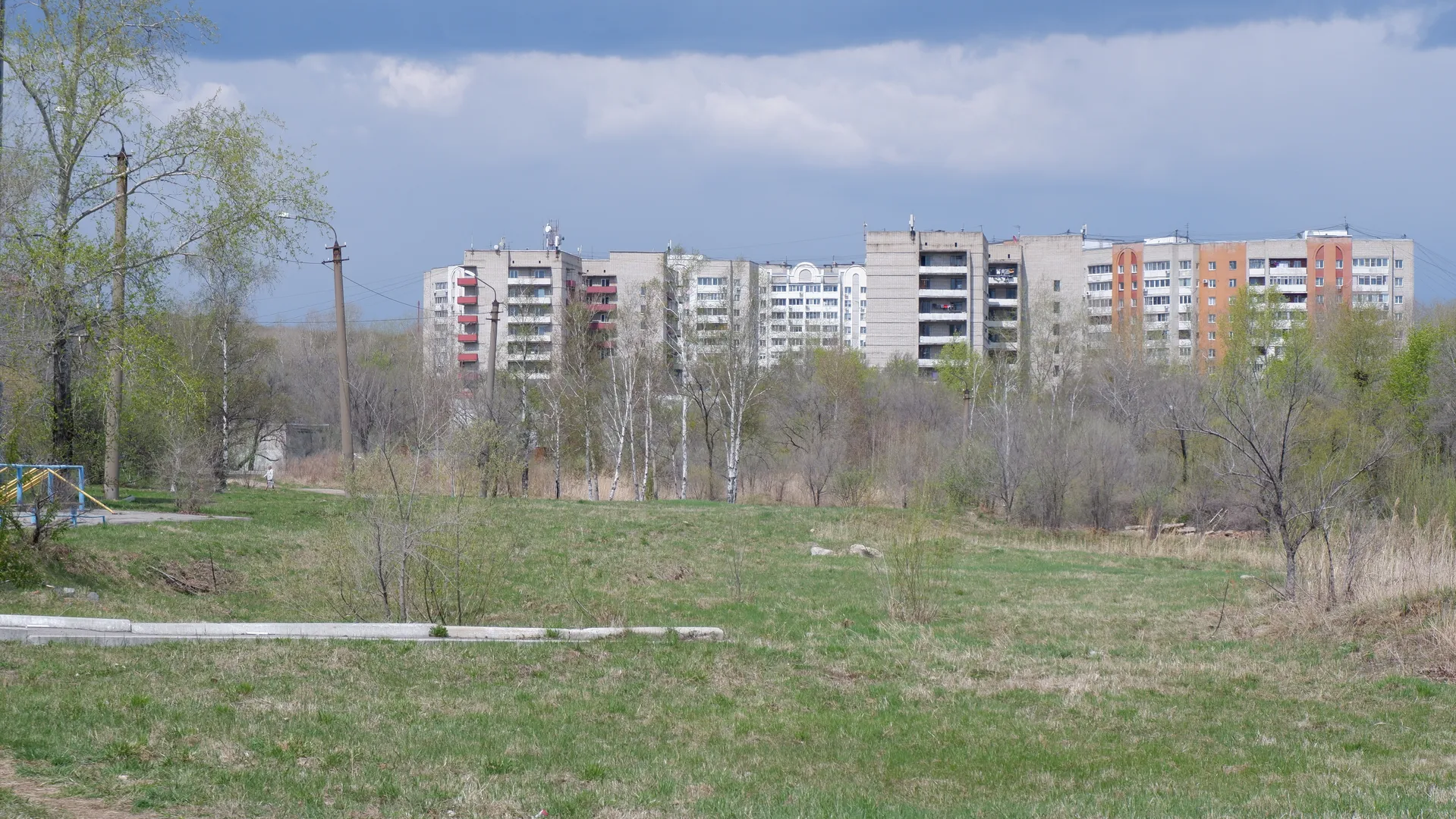 In front of a multi-story residential building is a meadow with scattered trees and shrubs. The building’s exterior walls are a mix of gray, orange, and white. The sky is covered with large clouds, and the weather looks overcast.