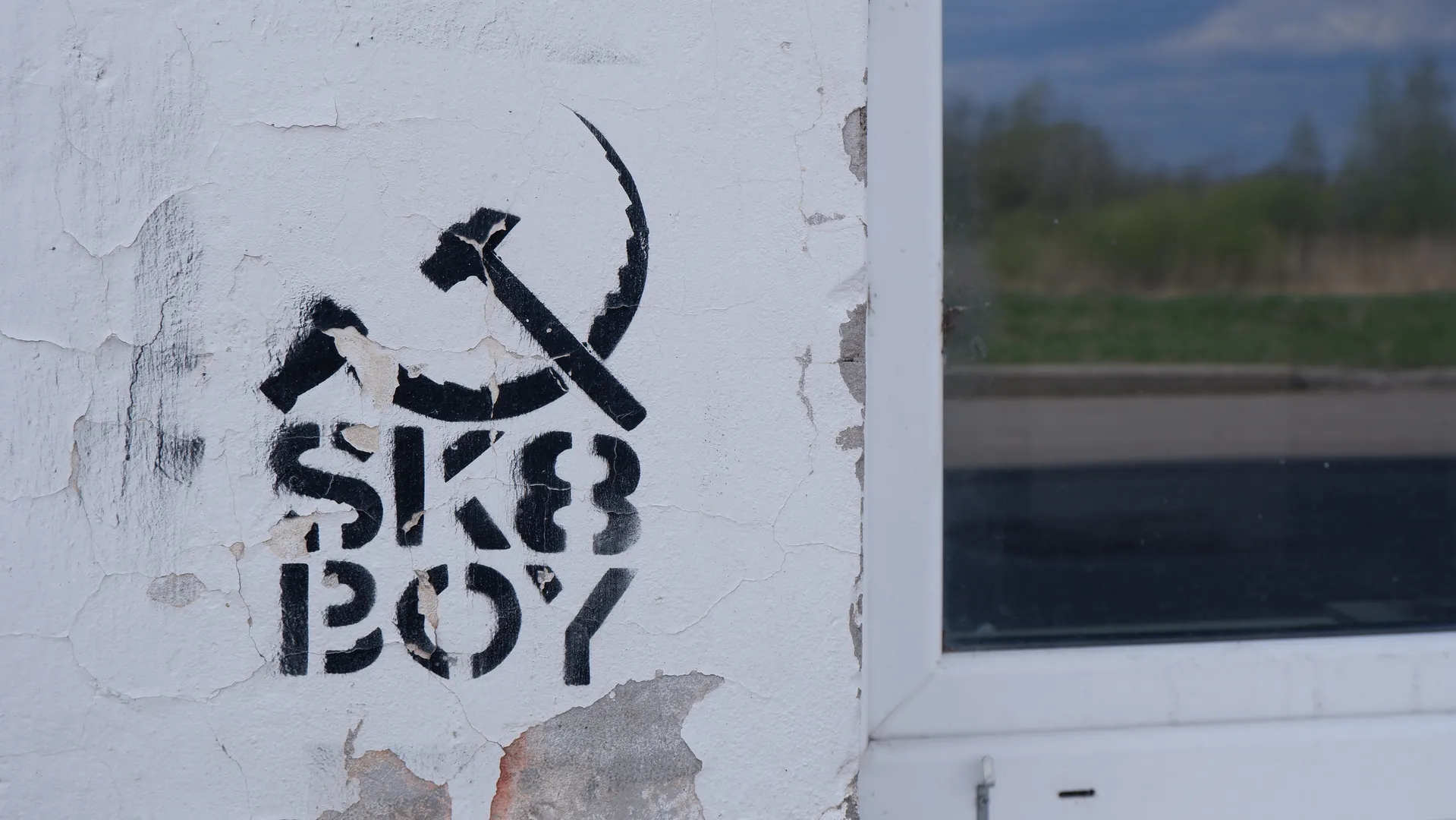 A black graffiti on the wall features a hammer and sickle symbol, with the text “SK8 BOY” underneath. The wall shows signs of peeling, indicating some wear and tear. In the background, a green meadow and trees can be seen.