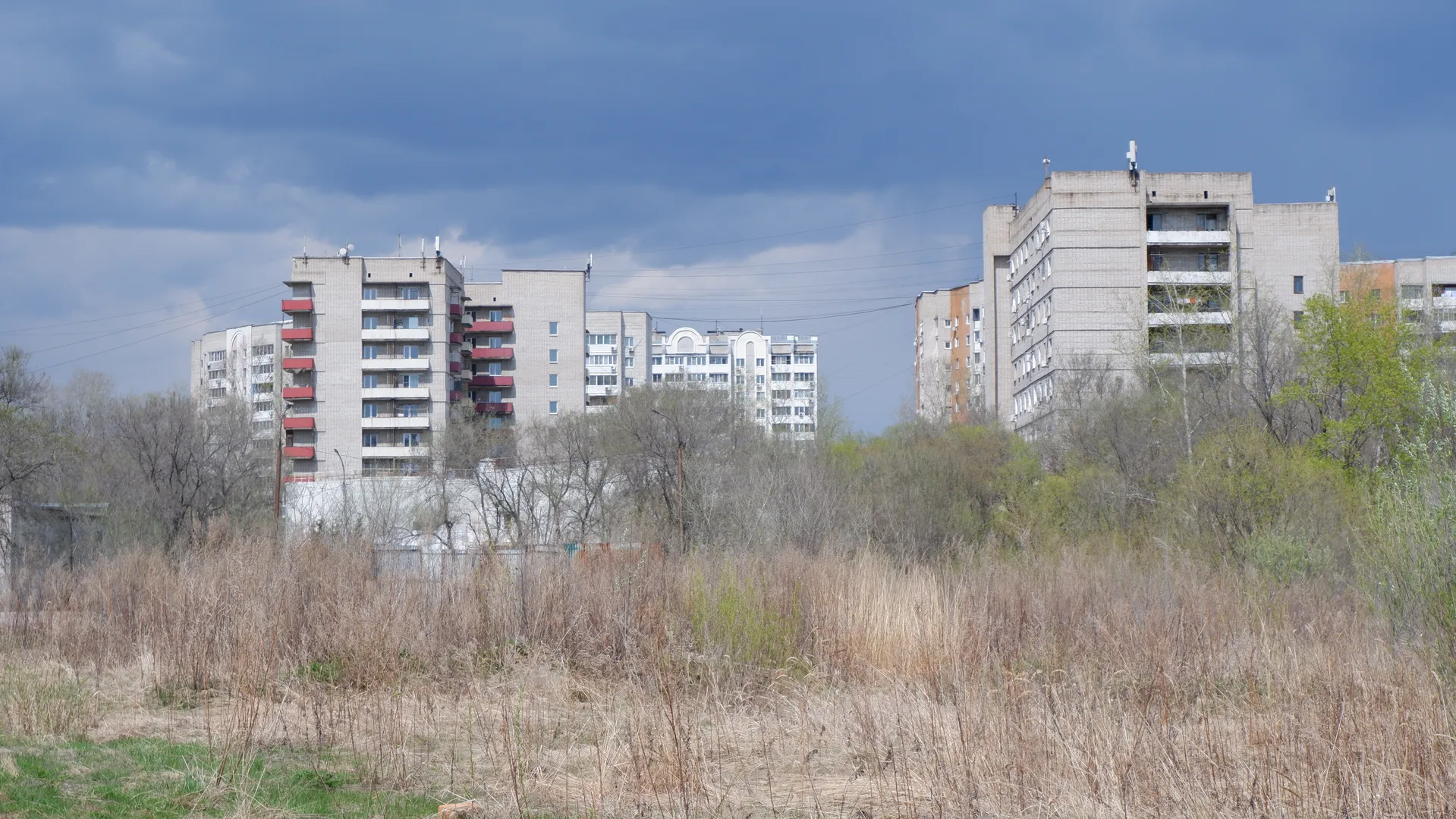 Behind the multi-story buildings are dense trees and shrubs. The buildings’ exterior walls are varied in color, with neatly arranged windows. The sky is partly covered with clouds, but sunlight still shines through.