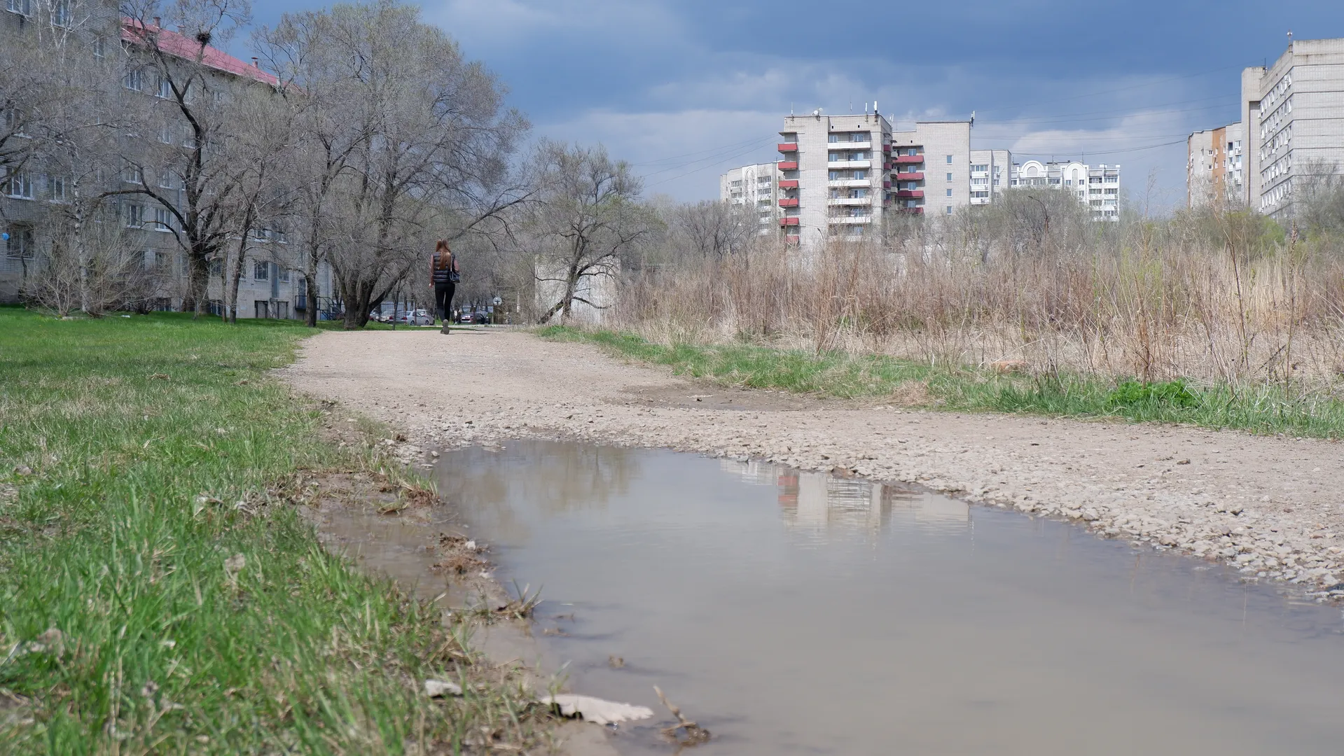 A dirt road runs through the meadow with a puddle on it. In the distance, a person is walking along the road, with multi-story buildings visible in the background. The sky has clouds, and the light is bright.