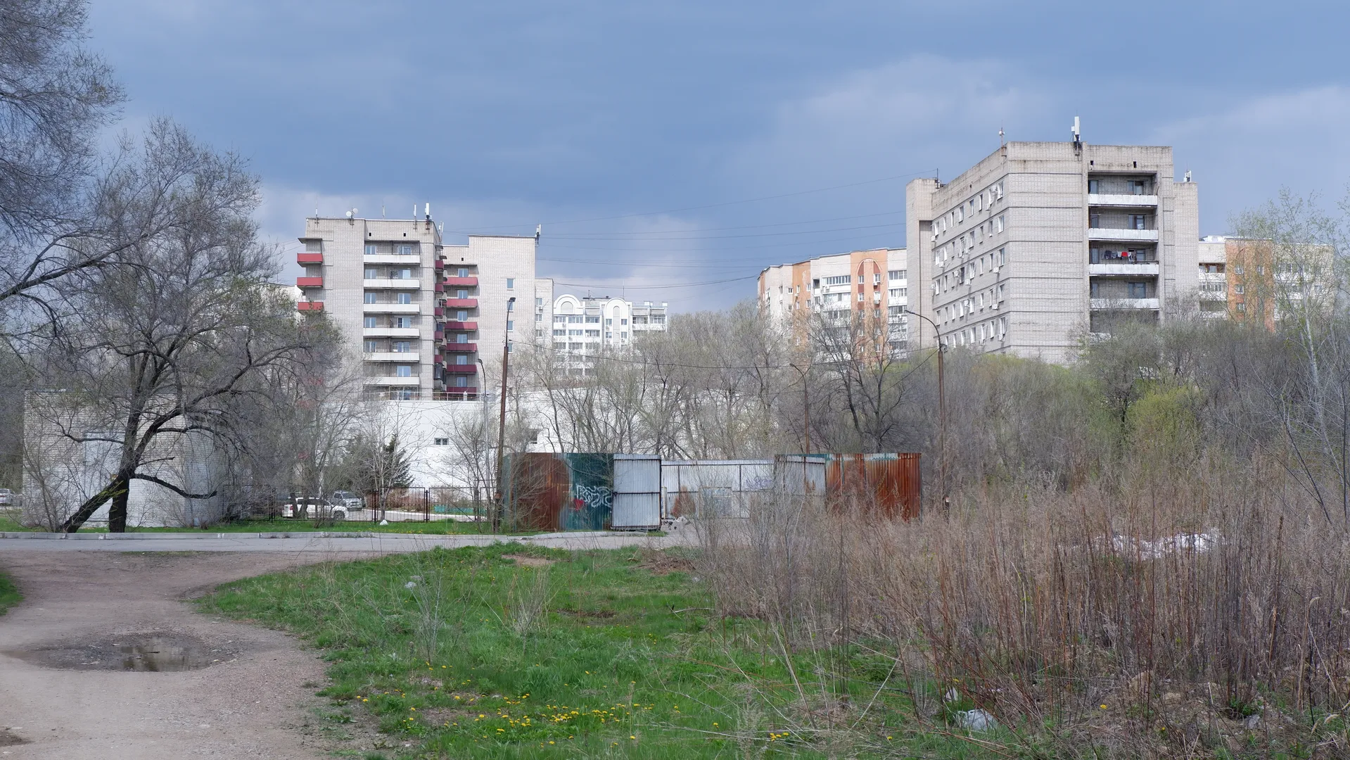 Behind the multi-story buildings are trees and shrubs, with a meadow in the foreground. The buildings’ exterior walls are varied in color, with neatly arranged windows. The sky has clouds, but the light is sufficient.