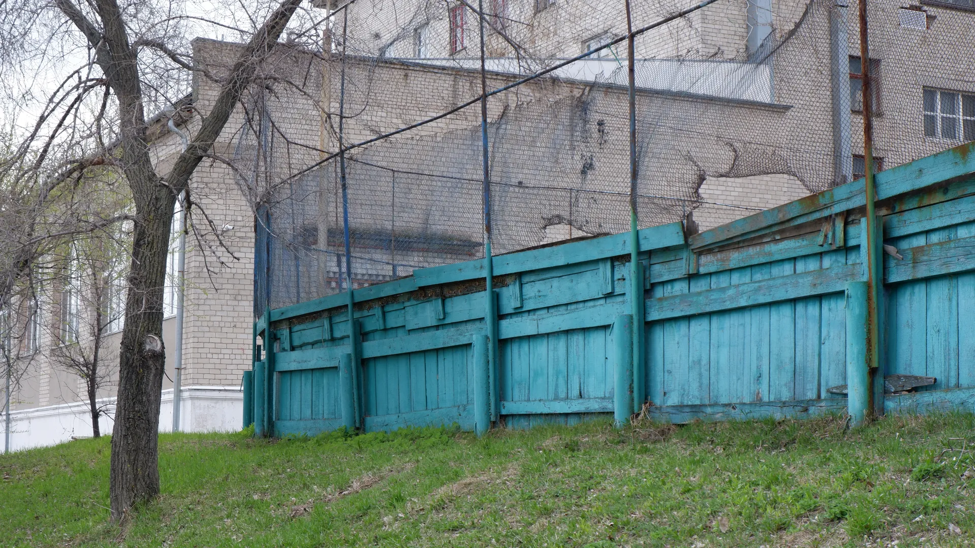 A blue wooden fence stands in front of a meadow, with a building behind it. The fence has barbed wire, and some areas are damaged. The building has a light-colored exterior with neatly arranged windows.