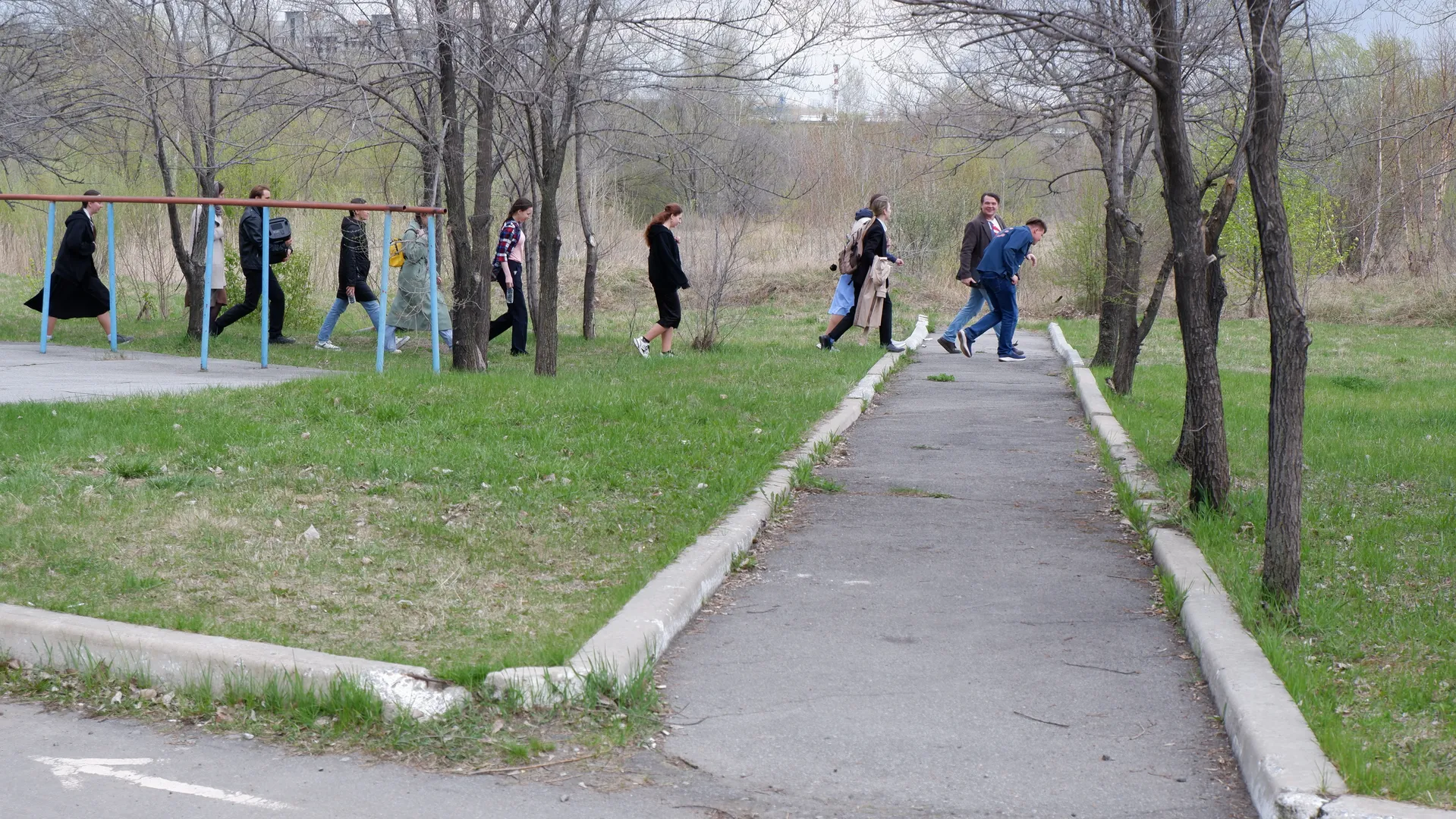 Several people are walking along a park path, with trees and grass beside them. In the distance, some people are standing next to a metal frame. In the background, there are buildings and open fields.