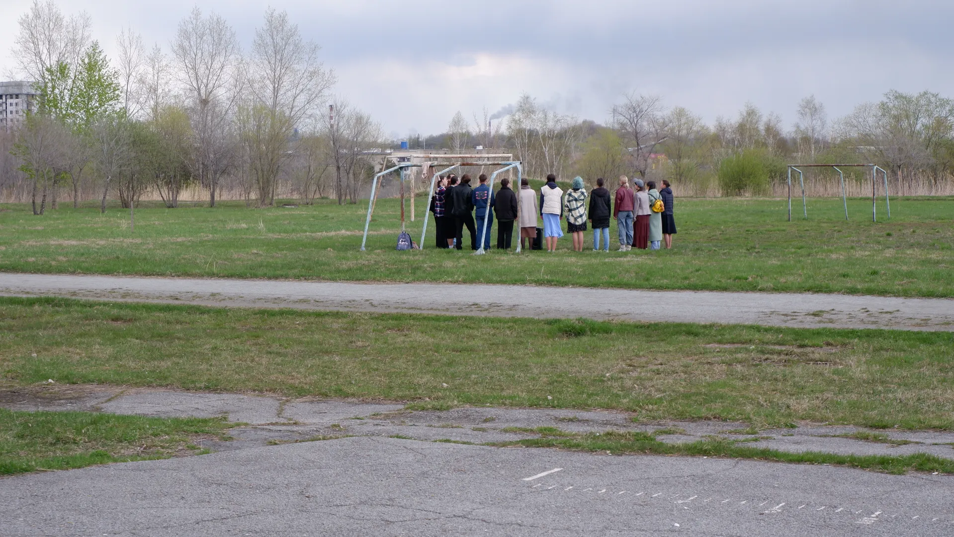 A group of people stands on the meadow, with their backs to the camera. A metal frame is in front of them, and the background shows open fields and trees. The sky is overcast, with buildings in the distance.