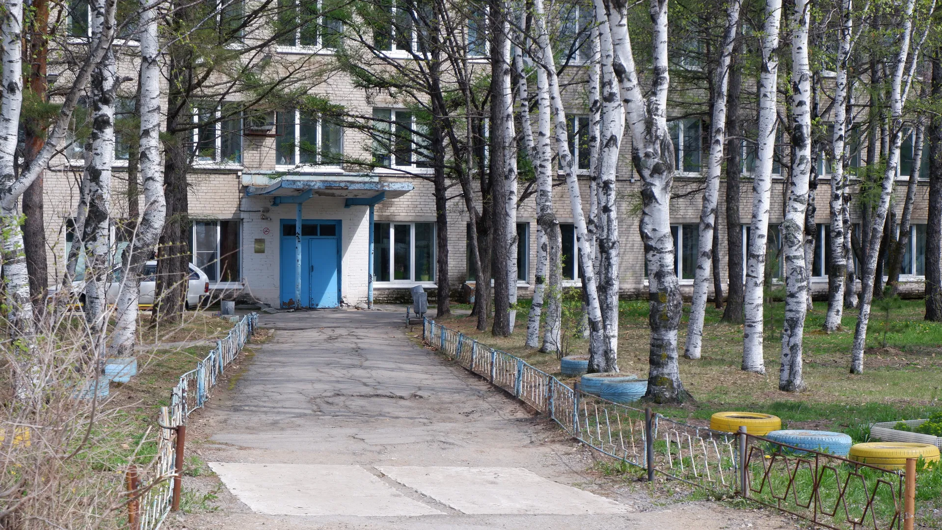 A path leads to the entrance of a building, where there is a blue door. On both sides are fences and trees, with some decorations on the ground. The building has a light-colored exterior and neatly arranged windows.