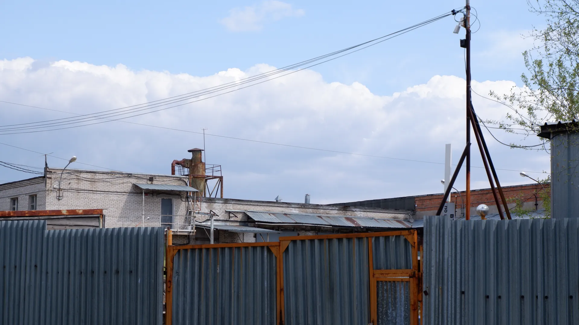 A tall iron sheet fence stands in front of an open iron gate. Behind the fence is a building with a chimney and antenna on the roof. The sky has white clouds, and power lines cross the frame.