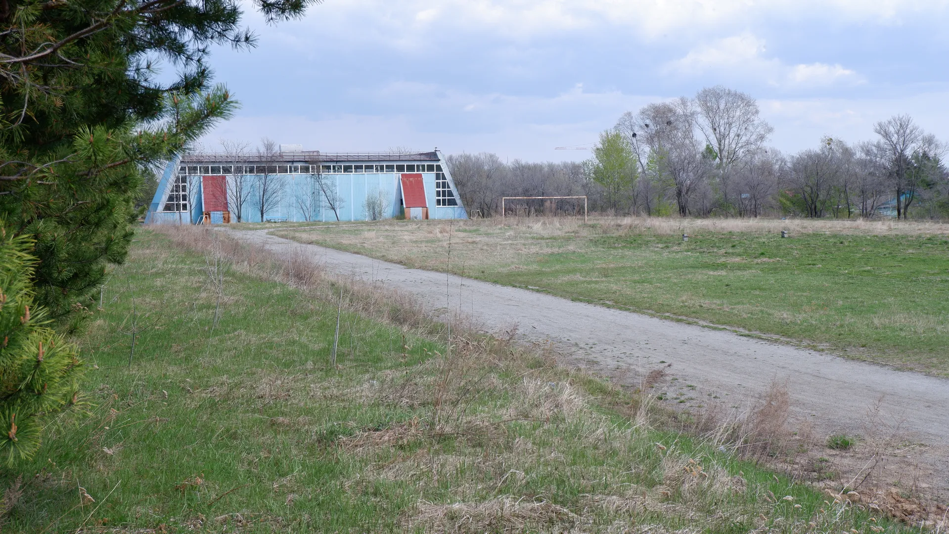 A path runs through the meadow leading to a blue building. The building has two red doors, surrounded by trees and shrubs. The sky has white clouds, and part of the meadow is dry.