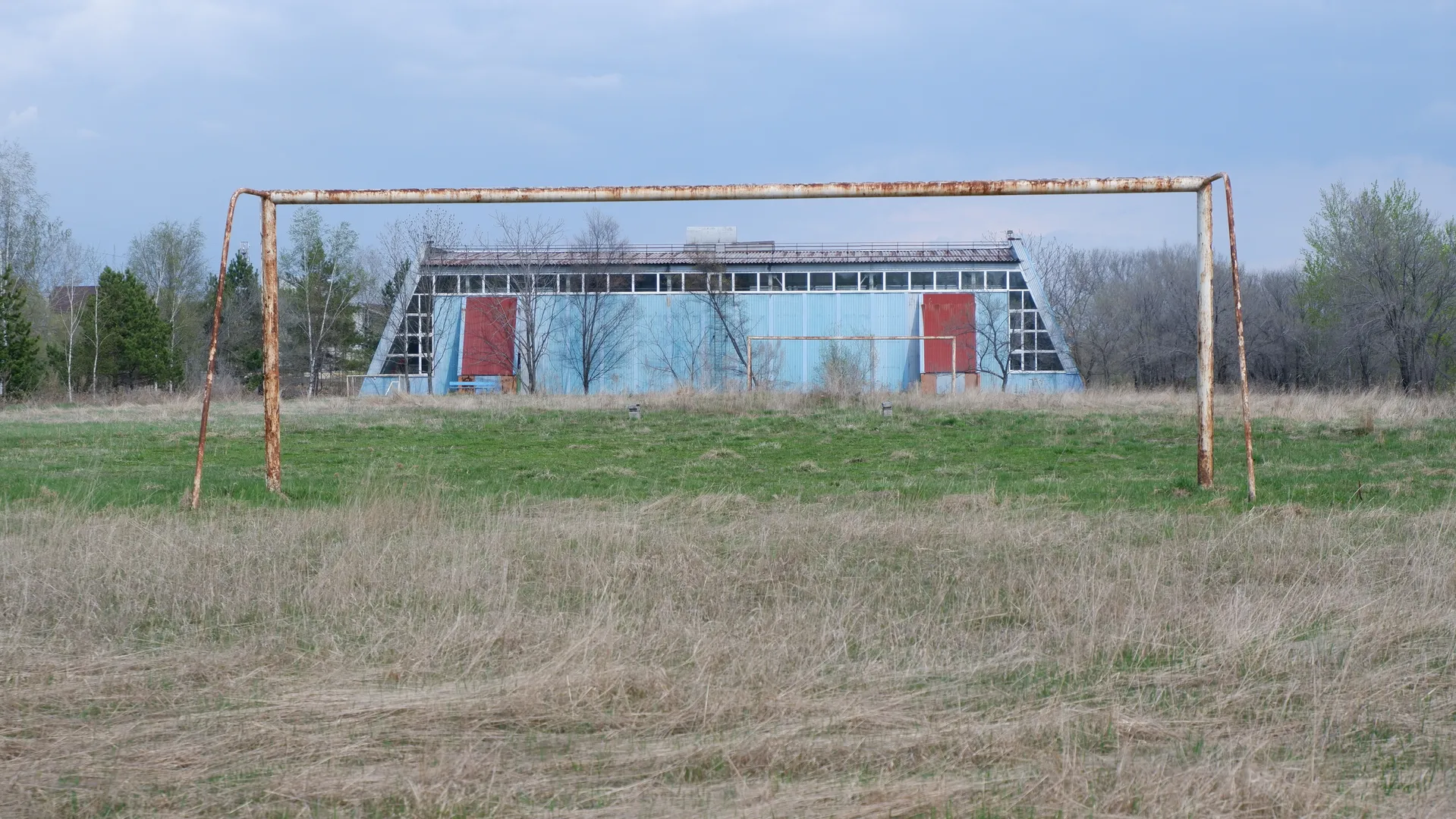 On a dry meadow, there is a rusty football goalpost. In the background, there is a blue building with two red doors. In the distance, trees and the sky are visible.