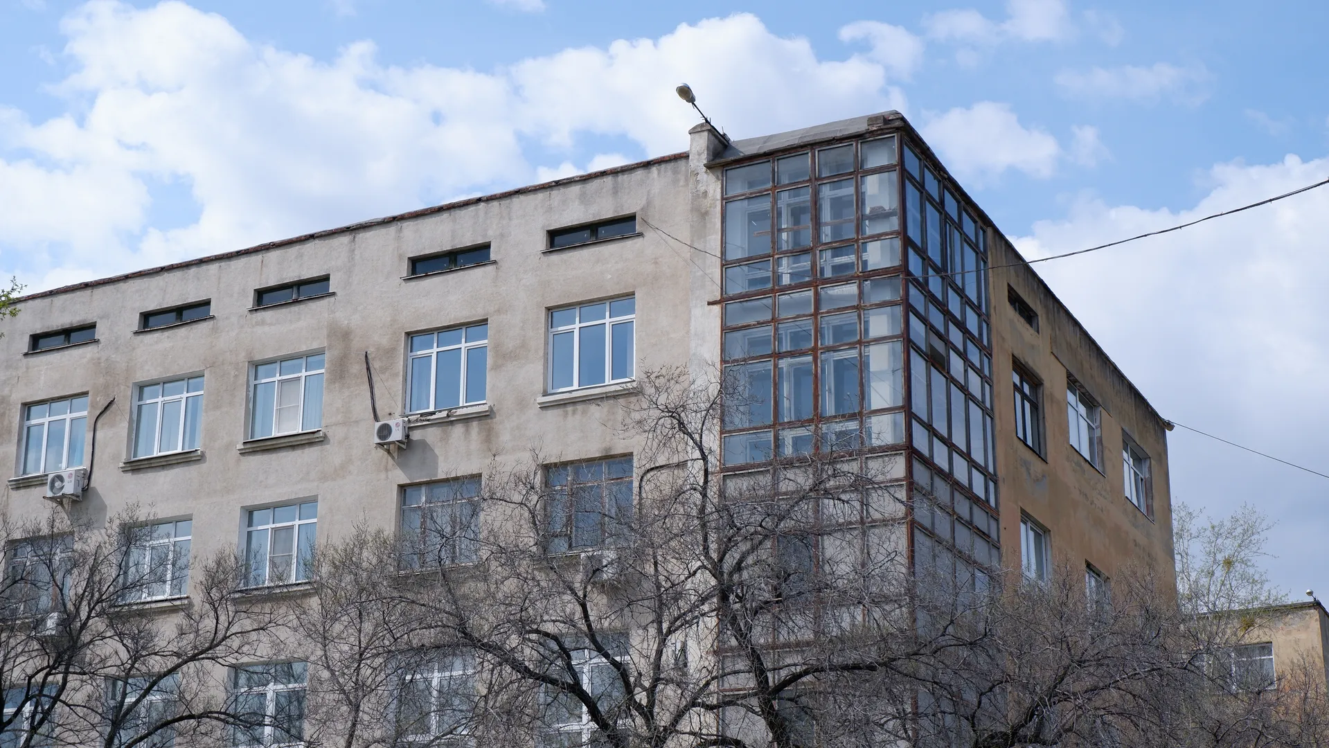 The exterior wall of a multi-story building is light-colored, with large glass windows at the corners. The top of the building has lighting, and the windows are neatly arranged. In the foreground, several trees with bare branches can be seen.