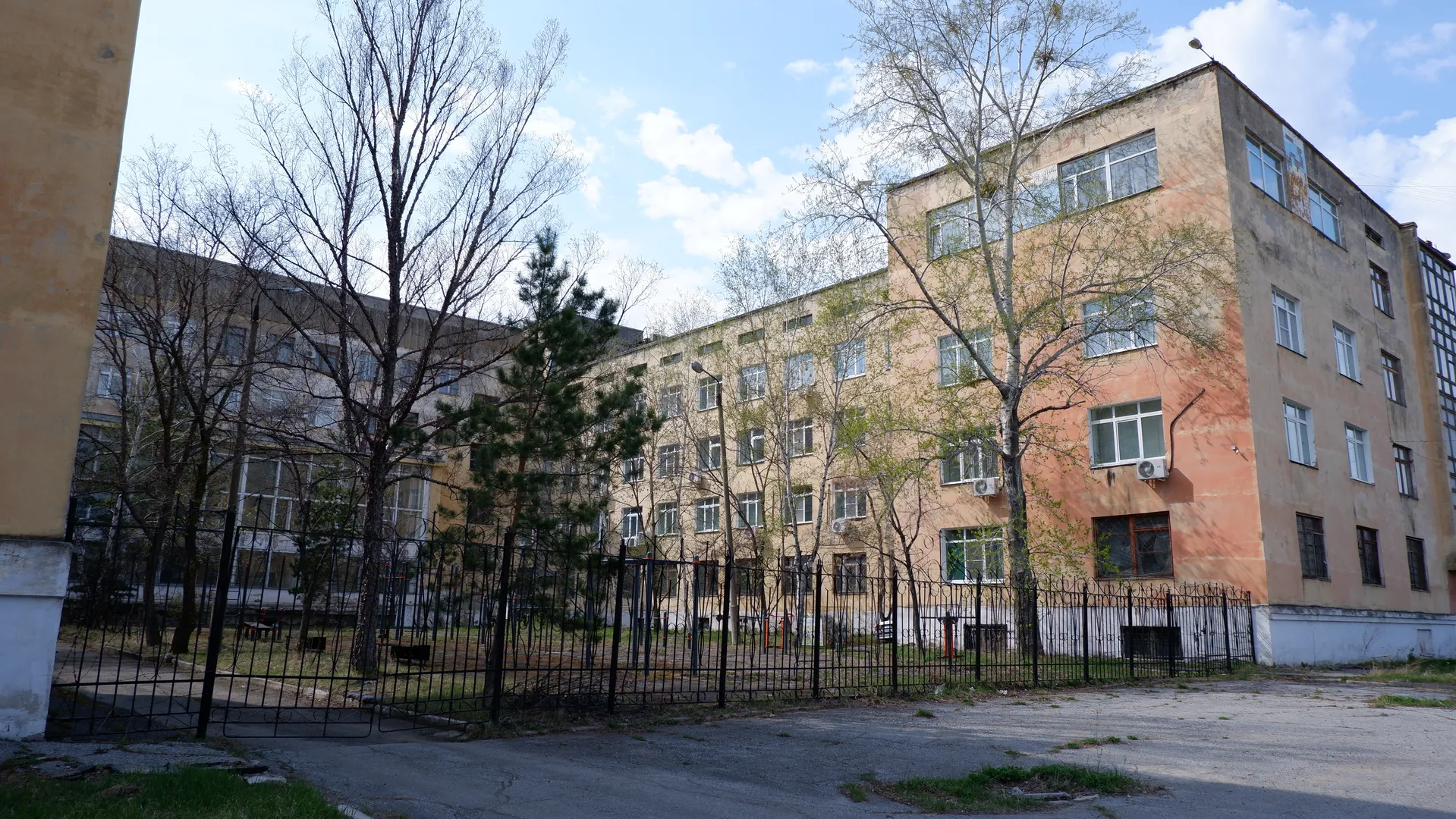 In front of a multi-story building, there are iron fences and several trees. The building has a light-colored exterior with neatly arranged windows. In the foreground, there is a small path made of cement.