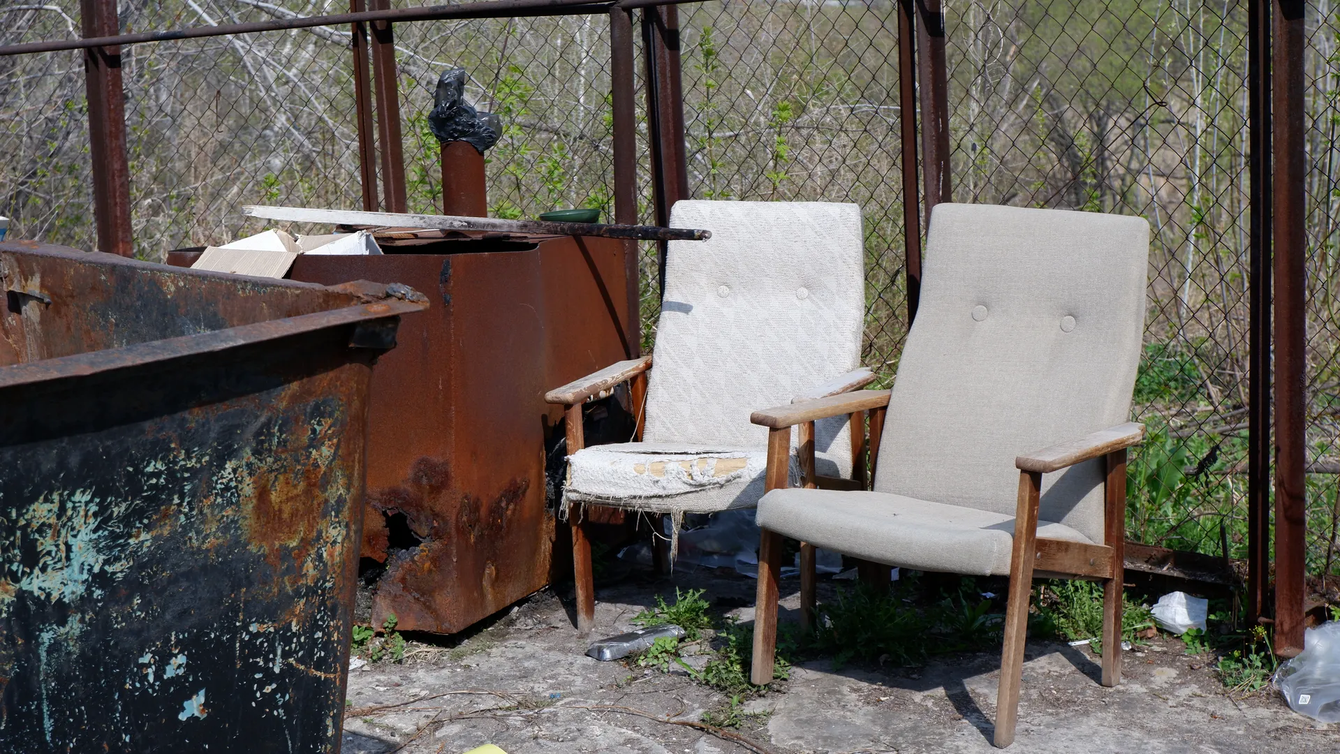 Two old chairs are placed in front of a wire fence, next to a rusty metal box. The box has some debris and a black plastic bag on top. In the background, there are trees and green plants.