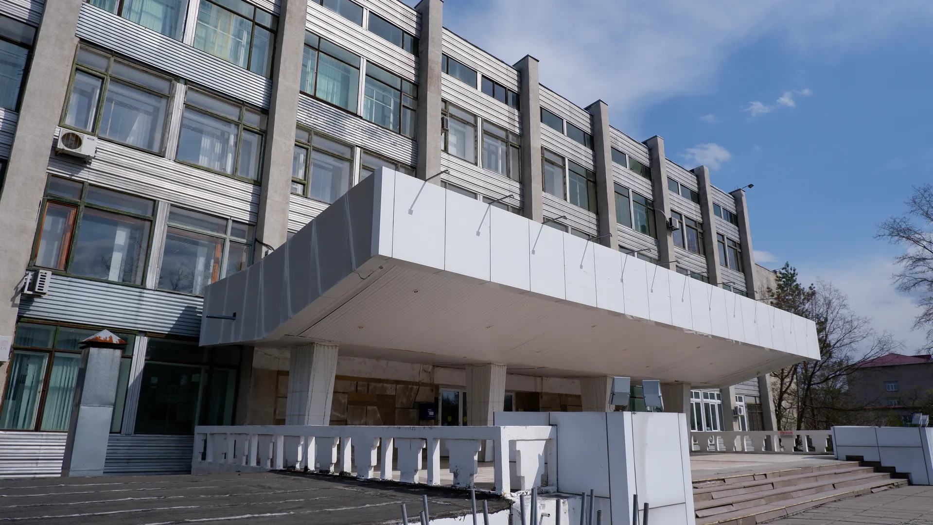 At the entrance of a modern building, there is a large sunshade. The building has multiple layers of windows and air conditioning units. In front of the entrance, there are stairs and a handrail, with a clear sky above.