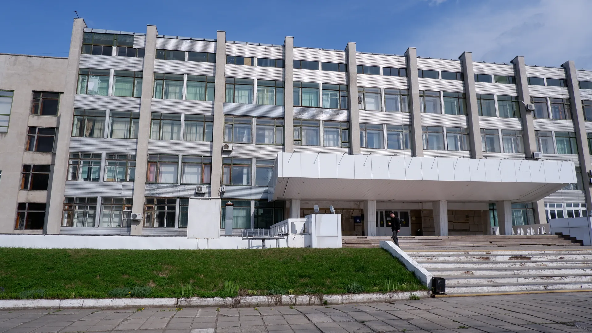 The front of a multi-story building with many windows and a light-colored exterior. There are stairs and a handrail in front of the building, with a person standing on the stairs. The sky is clear.